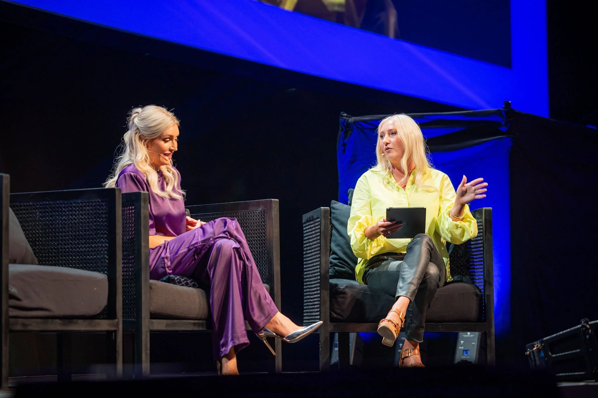 Two women are seated on a stage, engaged in conversation. The woman on the left has long blonde hair and is wearing a purple outfit, while the woman on the right has long blonde hair and is dressed in a yellow top and dark pants, holding a tablet.