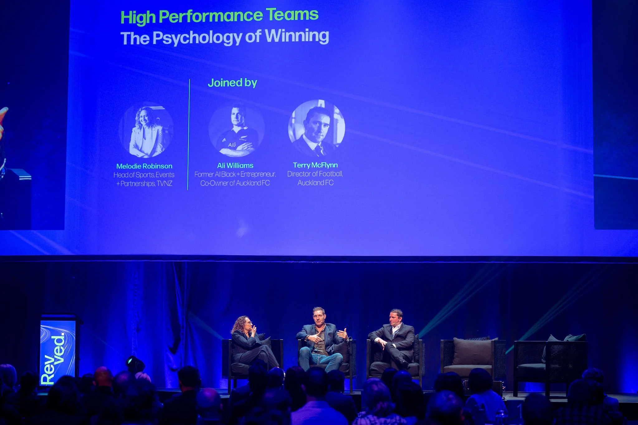 Panel discussion on stage at Revved with three people seated in chairs, behind a large screen displaying presentation slide that reads 'High Performance Teams: The Psychology of Winning,' along with photos and names of three presenters.