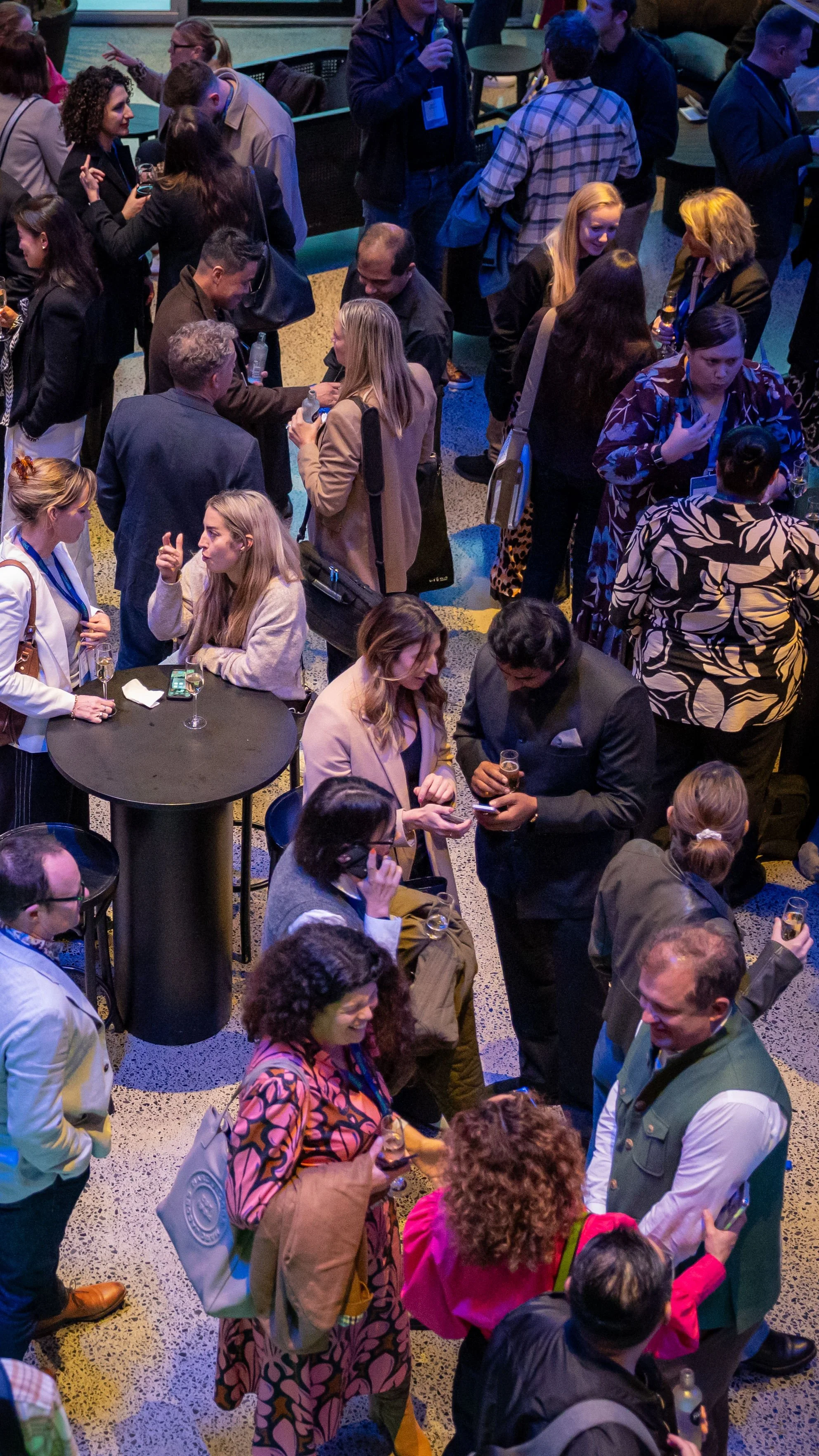 A crowded indoor networking event with people talking, drinking, and mingling around high tables and in groups, with some holding glasses of beverages.