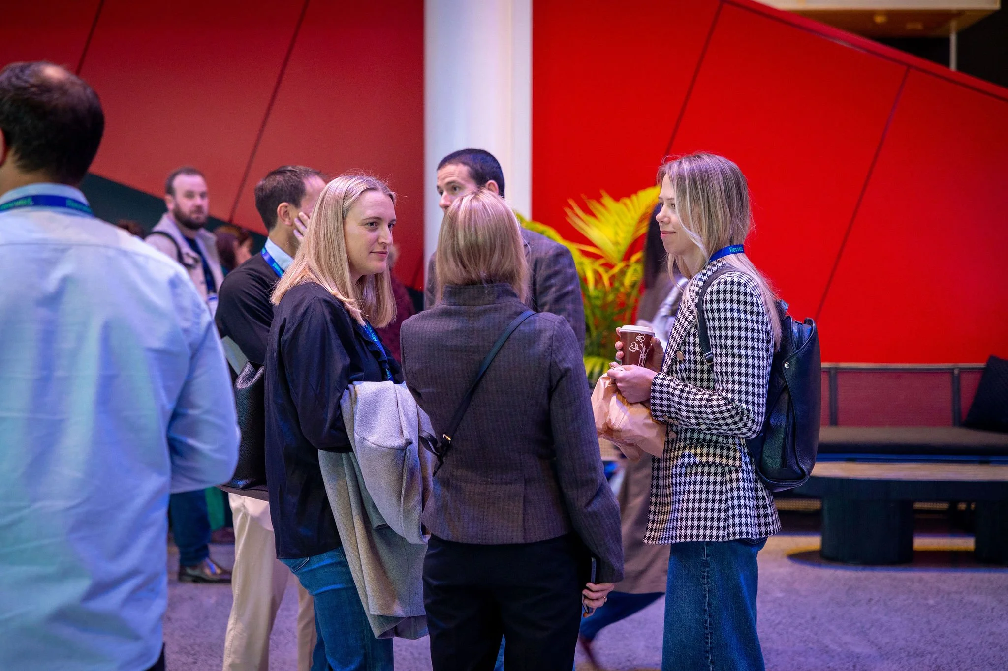 A group of people standing and talking in front of a red wall at an indoor event, with some holding coffee cups.