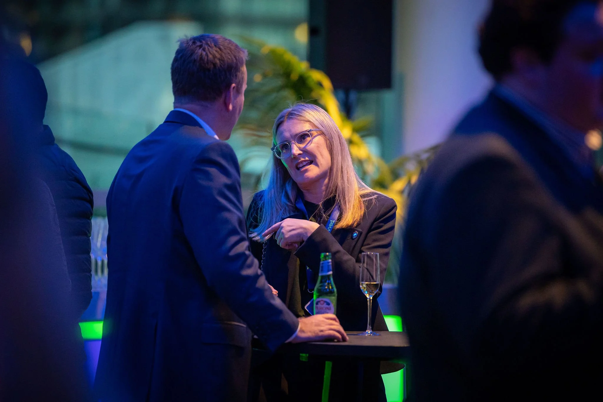 A group of people at a social event, with two individuals in conversation. One woman with long blonde hair and glasses is talking to a man with short hair, both dressed in dark jackets. There are drinks on the table, including a beer bottle and a gla