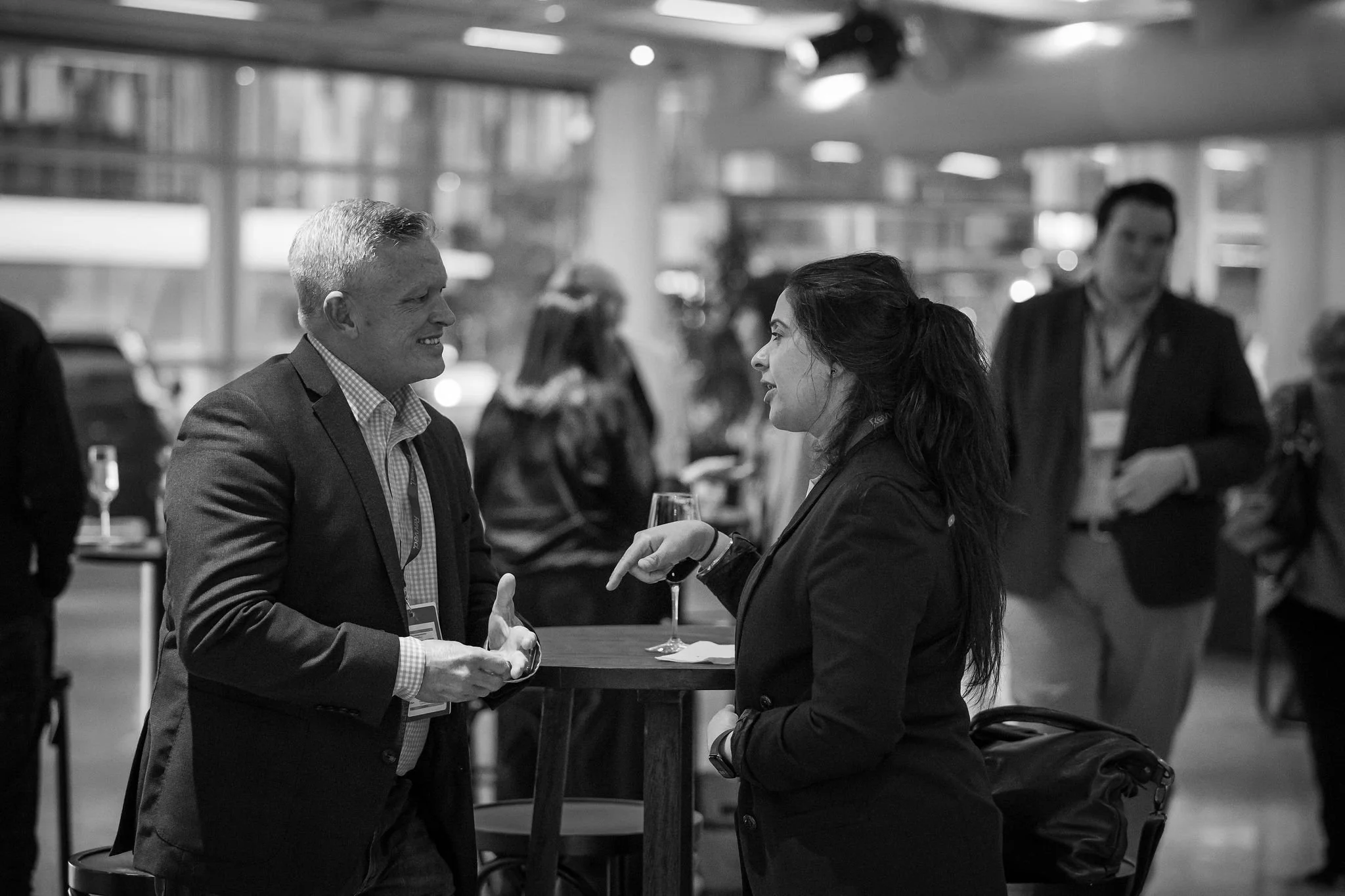 Two people, a man and a woman, are engaged in a conversation at a networking event or conference. The man is wearing a suit and is smiling, while the woman is holding a glass of wine and is talking to him. The background shows a busy indoor setting w