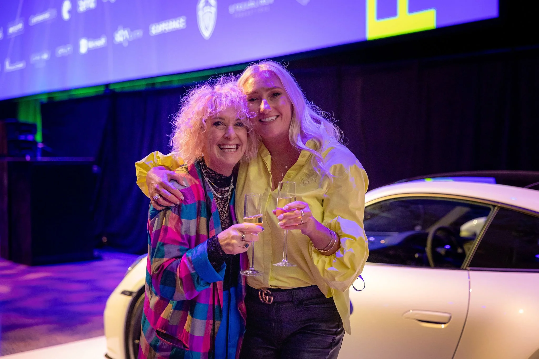 Two women are smiling and holding glasses of champagne at a celebration event. One woman has curly blonde hair and is wearing a colorful checkered blazer, while the other has long blonde hair and is dressed in a yellow shirt. They are standing in fro