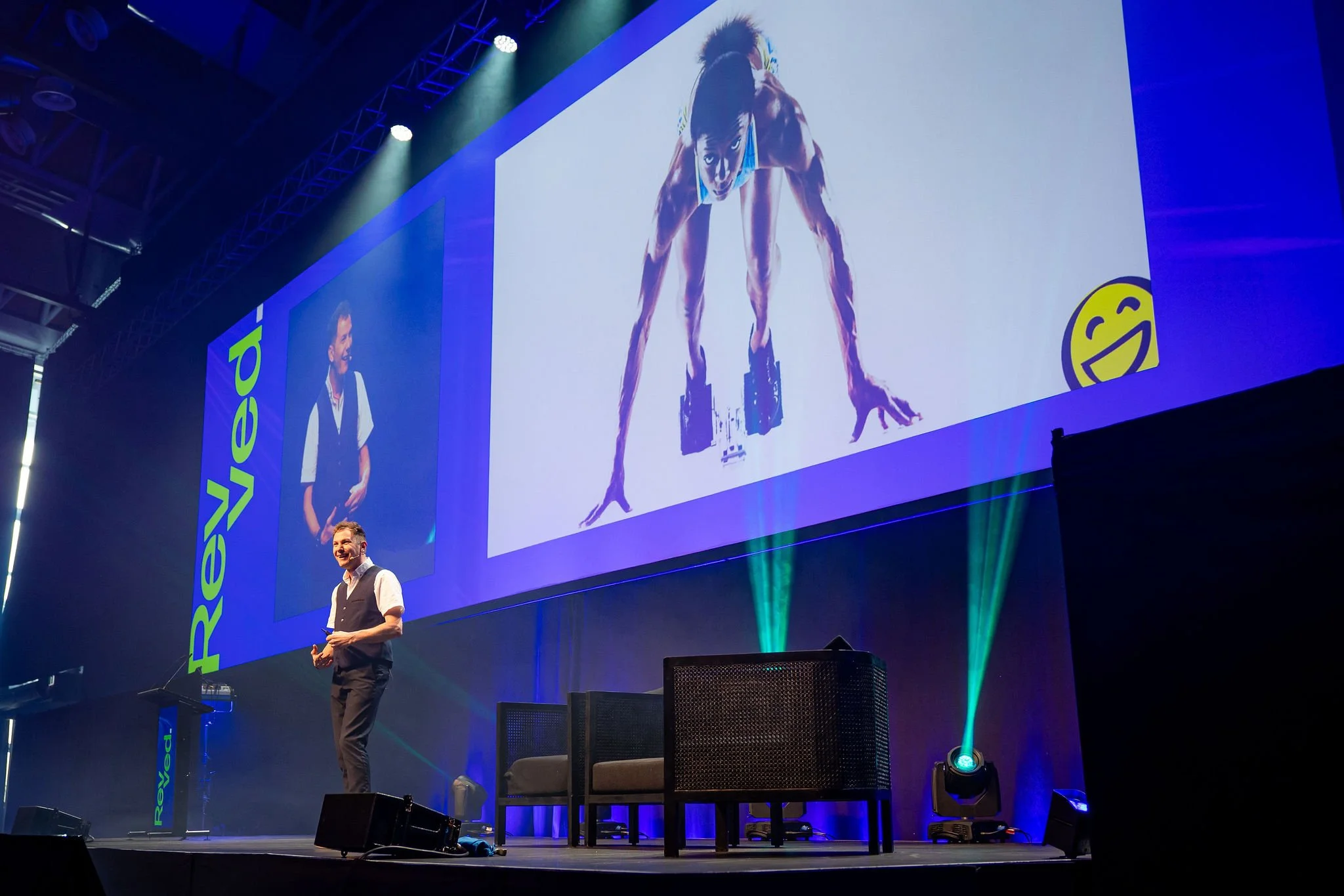 Keynote speaker on stage at Revved , with large screens behind him displaying a woman in a workout pose and a large yellow smiley face logo in the bottom right corner of the screen. The stage has blue and green lighting.