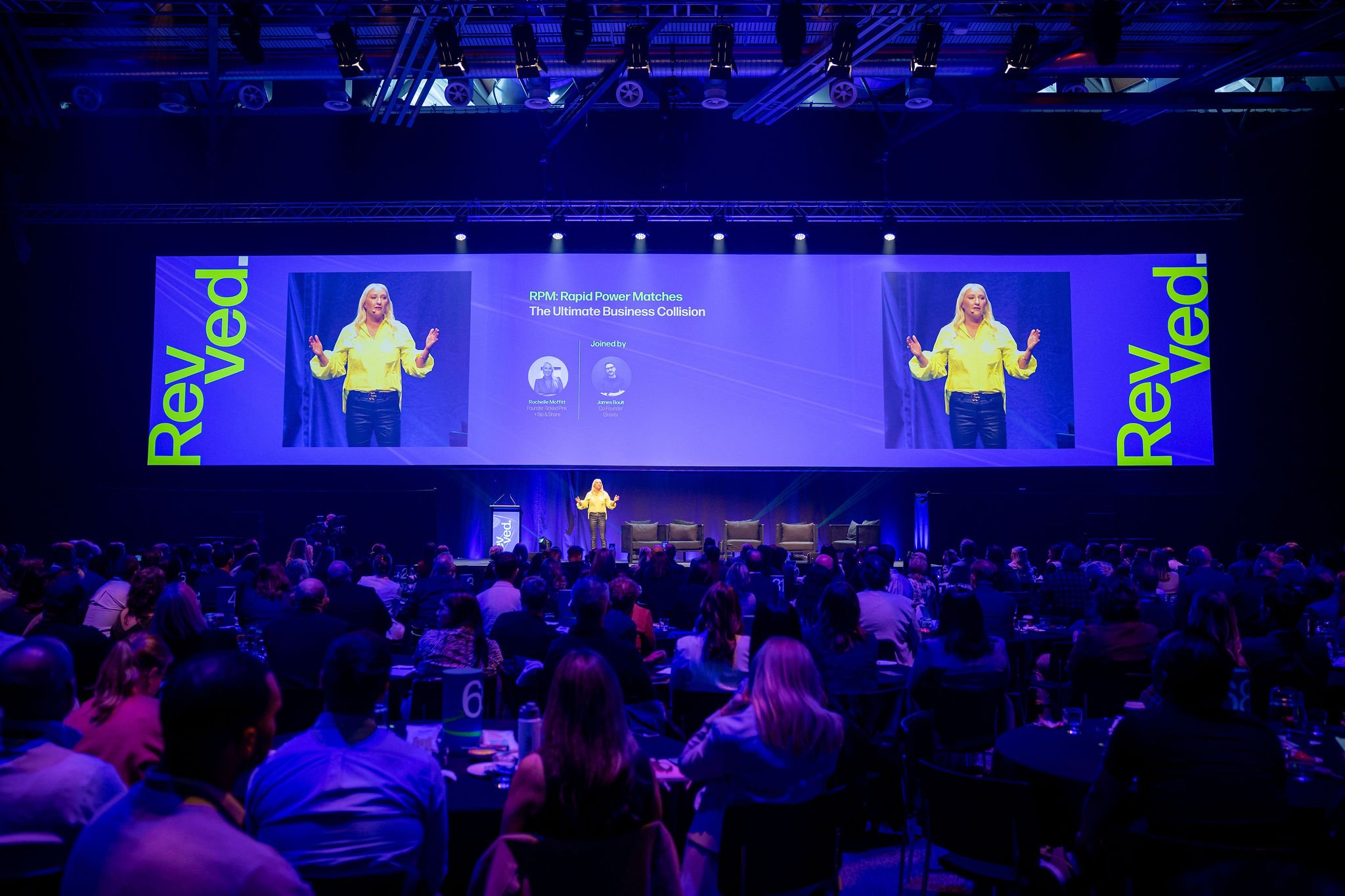 A woman in a yellow shirt giving a presentation on stage at a conference titled 'RPM: Rapid Power Matches The Ultimate Business Collision,' with an audience seated facing the stage in a dark auditorium.