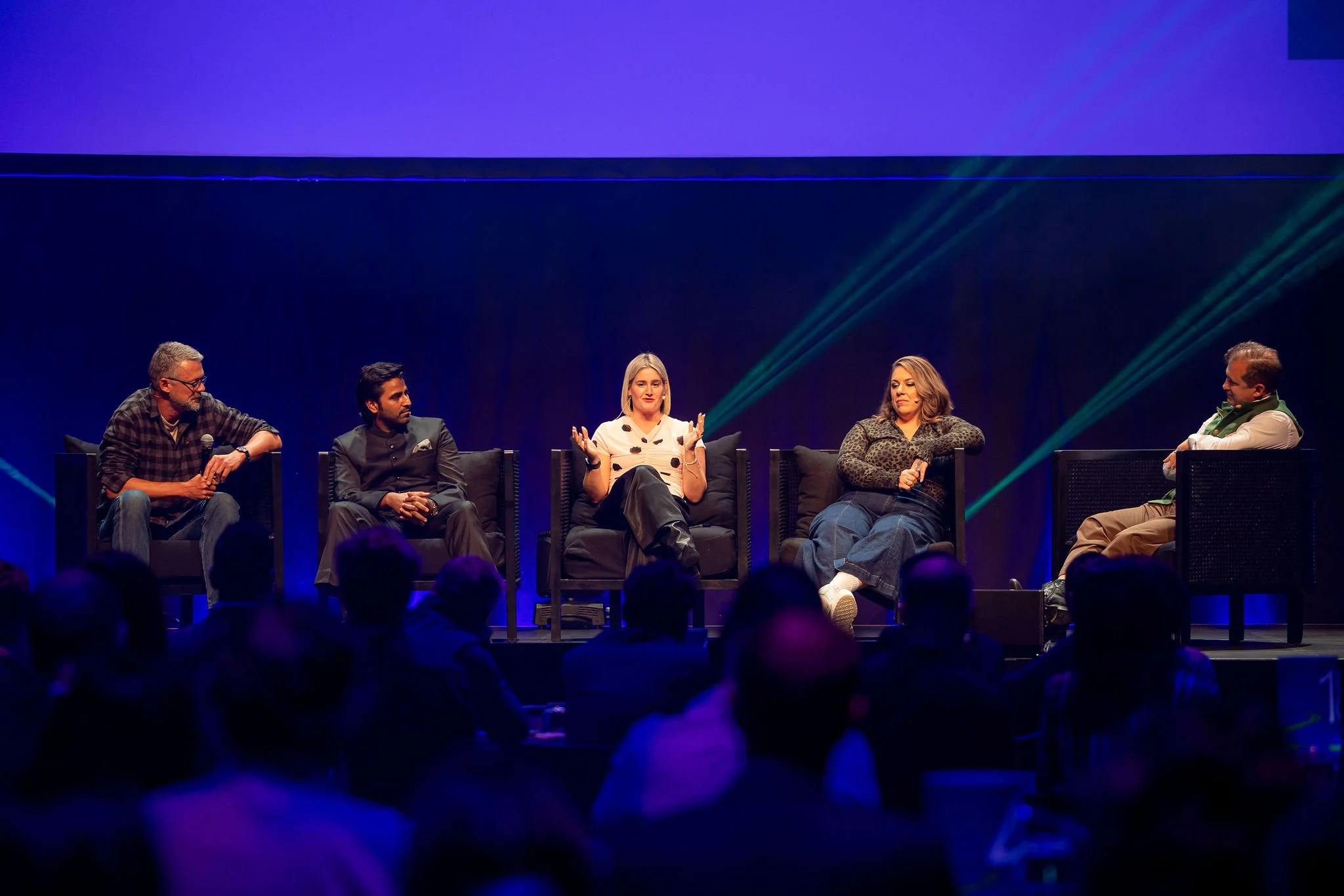 A Revved panel discussion with five people seated on stage in front of an audience, under purple and green lighting.