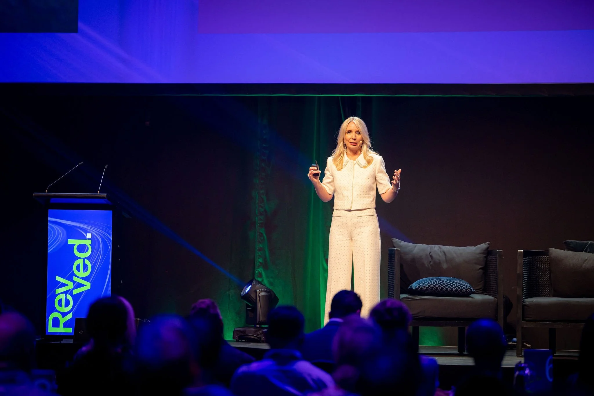 A woman with blonde hair giving a presentation on stage at a conference, with a large screen behind her displaying the word 'Revel.' She is wearing a cream-colored outfit and holding a remote control, with a microphone attached to her face.