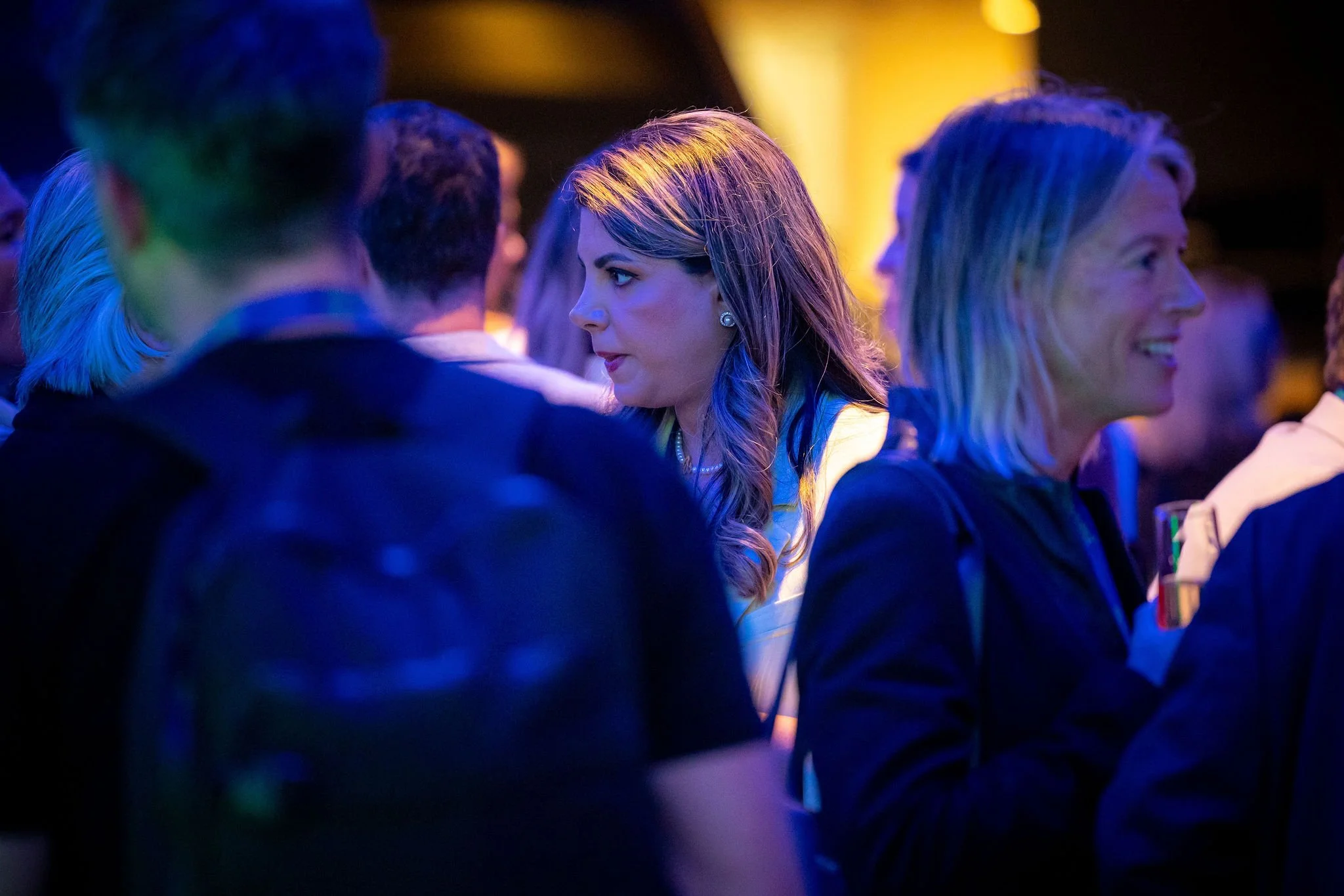 People at a social event, with a woman with long blonde hair in focus, illuminated by colored lights.