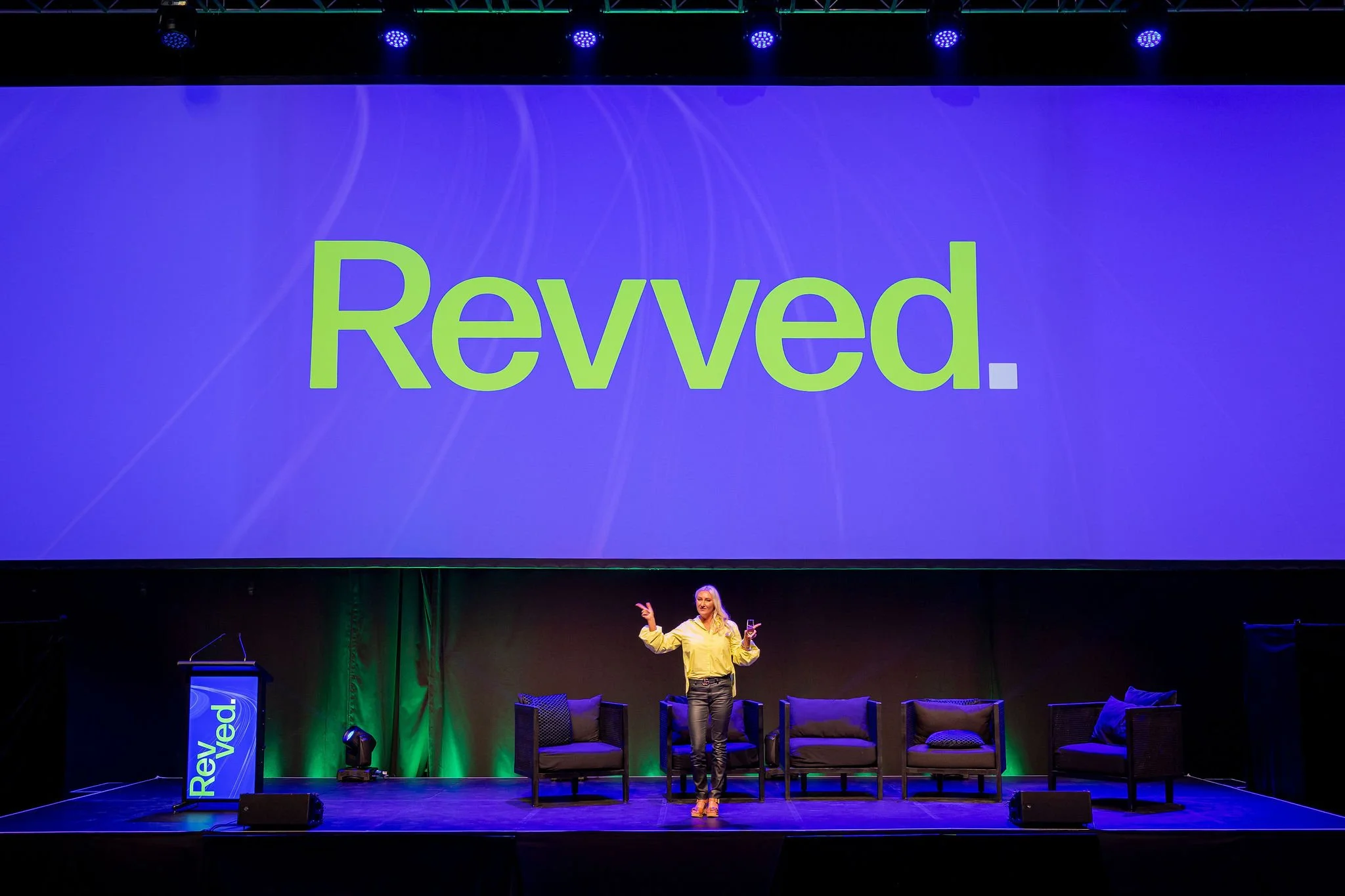 A woman on stage delivering a presentation at a conference, with a large screen behind her displaying the word 'Revoked' in green text on a blue background.