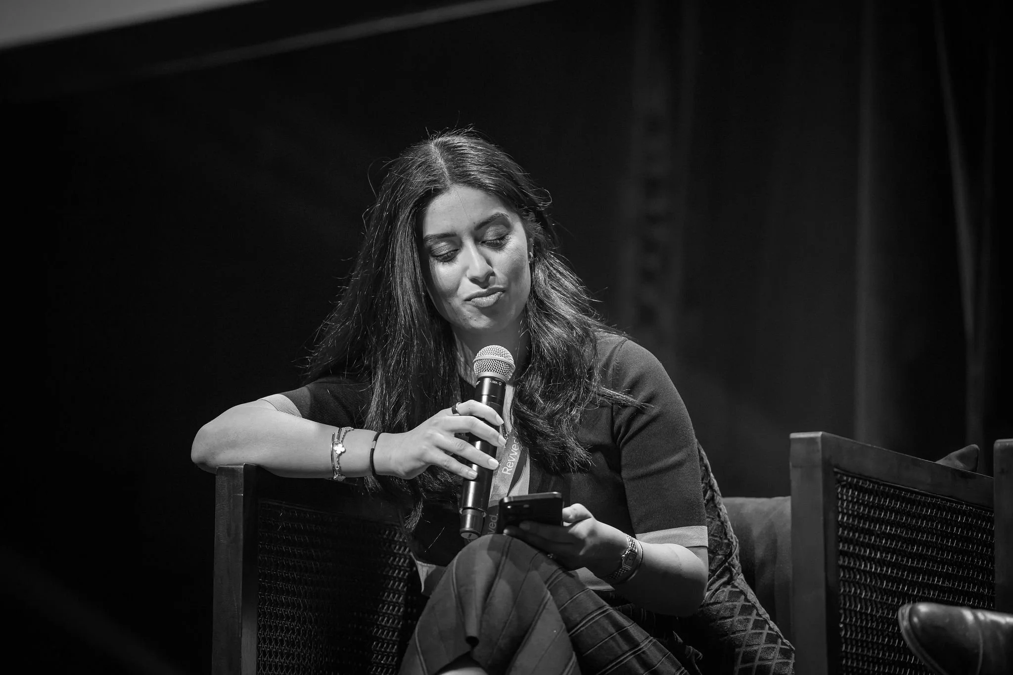 A woman with long dark hair and closed eyes, holding a microphone and looking down, sitting on a chair with a dark background.
