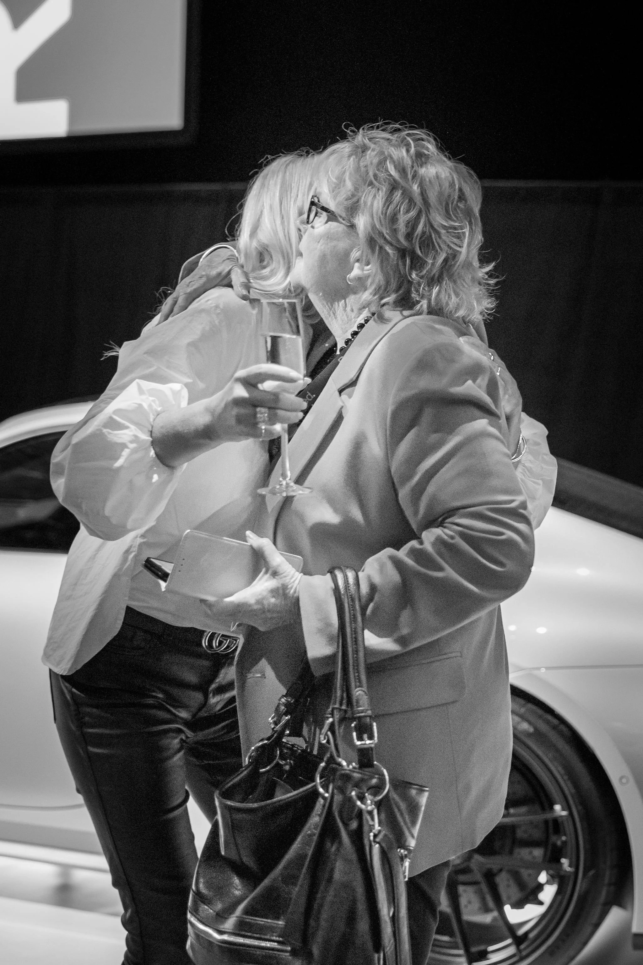 Two women embrace in a hug inside a setting with a car and a large screen in the background. One woman is holding a glass of champagne.