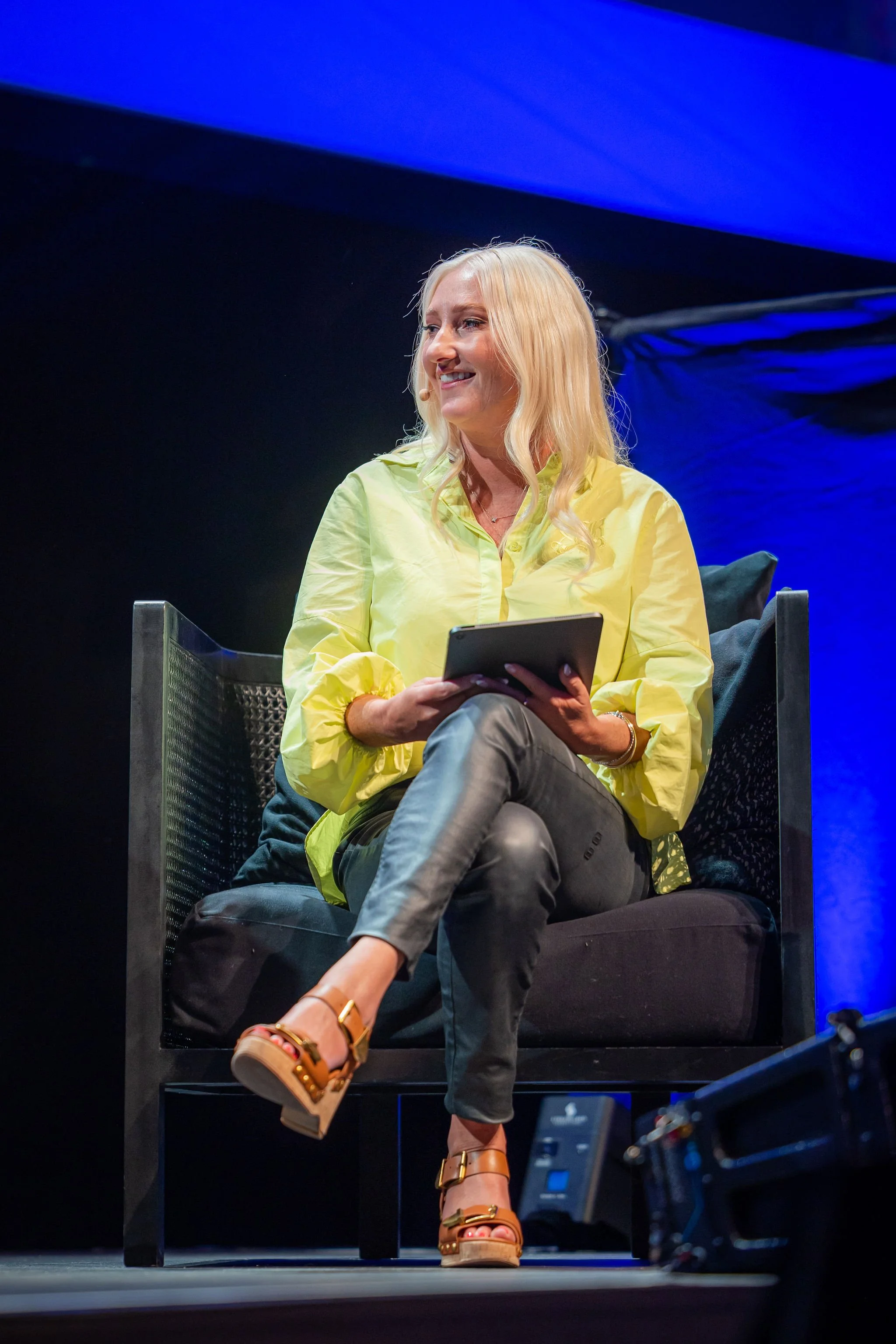 A woman with long blonde hair sitting on a black chair, smiling and holding a tablet, wearing a yellow shirt and high-heeled sandals, with stage lighting in the background.