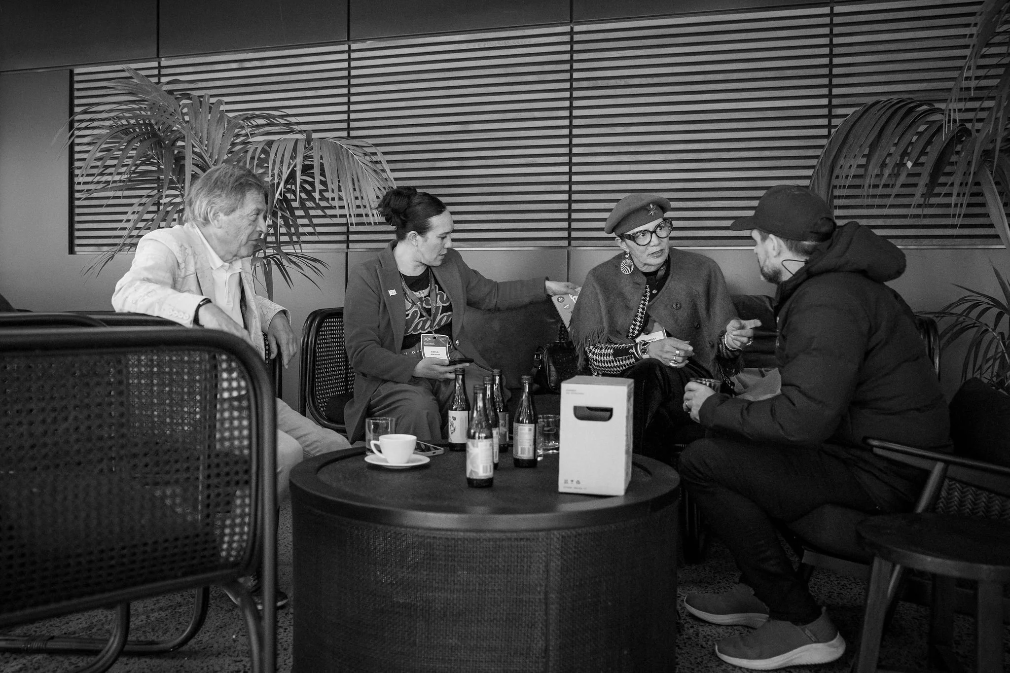 Four people seated around a table engaged in conversation in an indoor setting with wooden wall panels and potted plants.