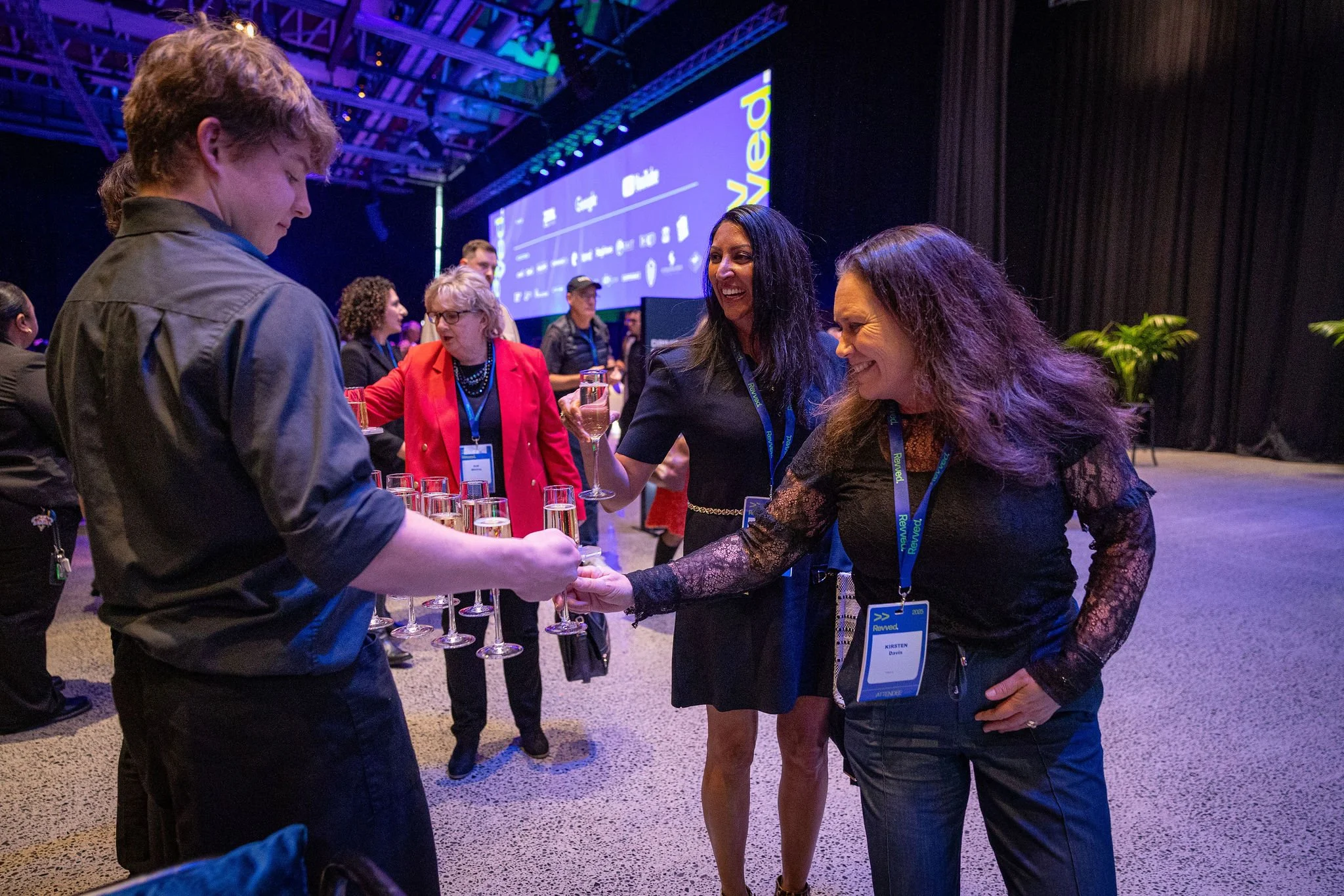 A group of well-dressed people at a conference or networking event, engaging in conversation and laughter. One woman in a black lace dress shakes hands with a young man, while others chat nearby. The background features a large screen with colorful g