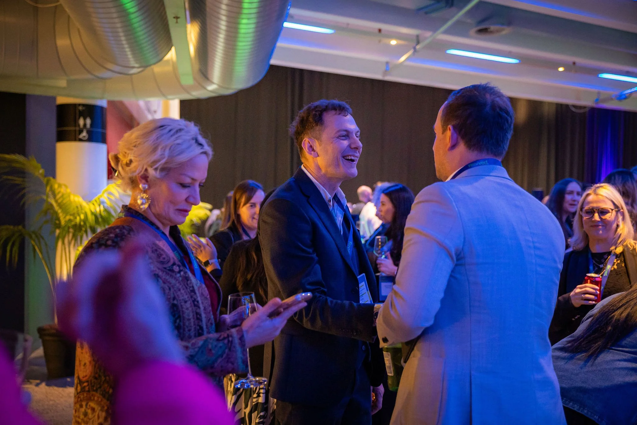 People attending a social event or conference, engaging in conversations, with a man smiling and talking to another man, surrounded by others holding drinks, under colorful lighting in a large indoor venue.