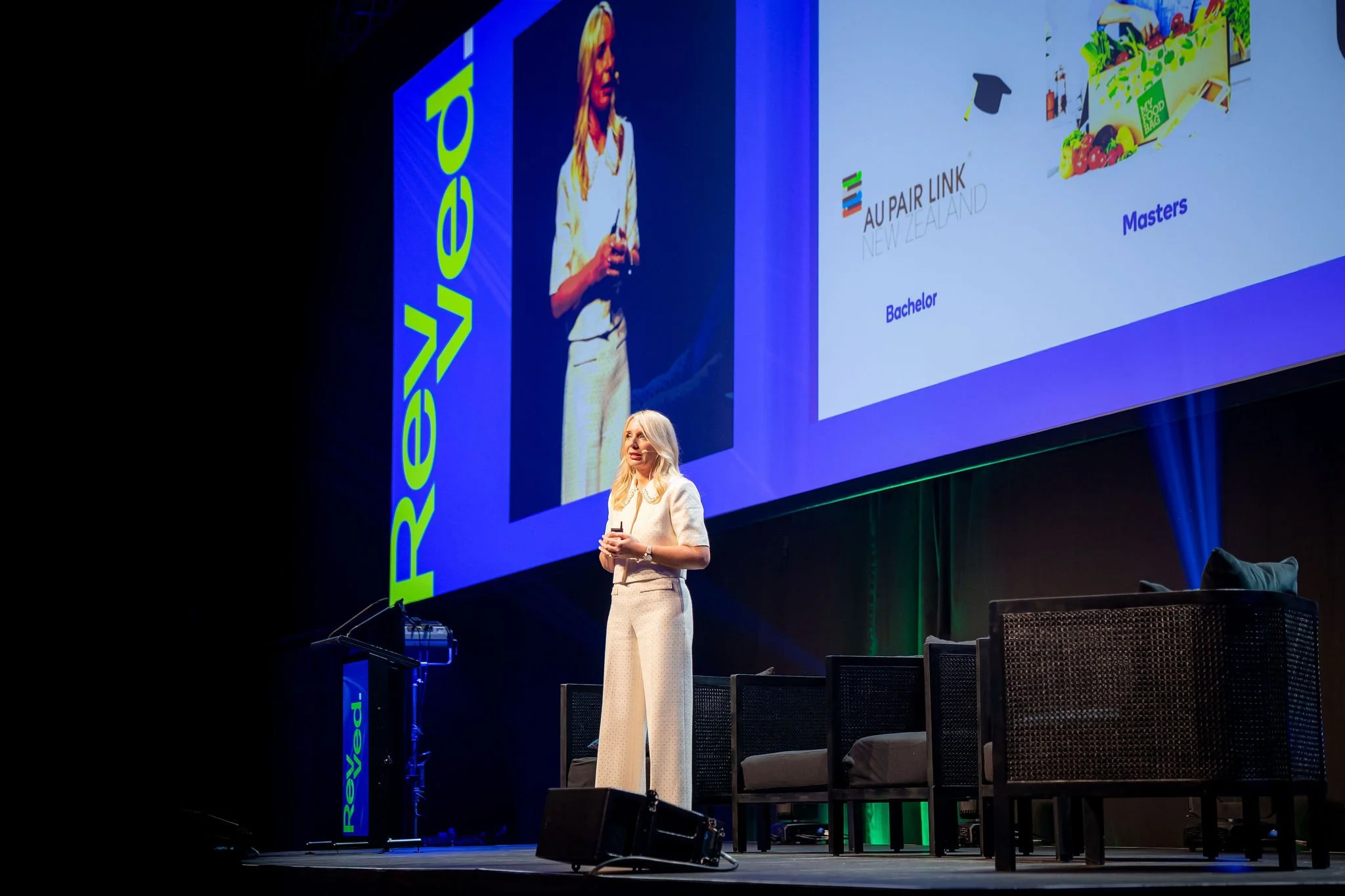 A woman speaking on stage at a conference or event. She is dressed in a light-colored suit and holding a microphone. A large screen behind her displays a presentation slide with logos and text, including the words "AU PAIR LINK NEW ZEALAND," "Masters