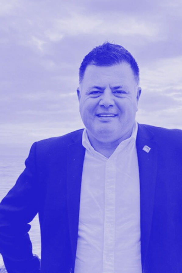 A man in a white shirt and dark blazer standing outdoors with a cloudy sky in the background.