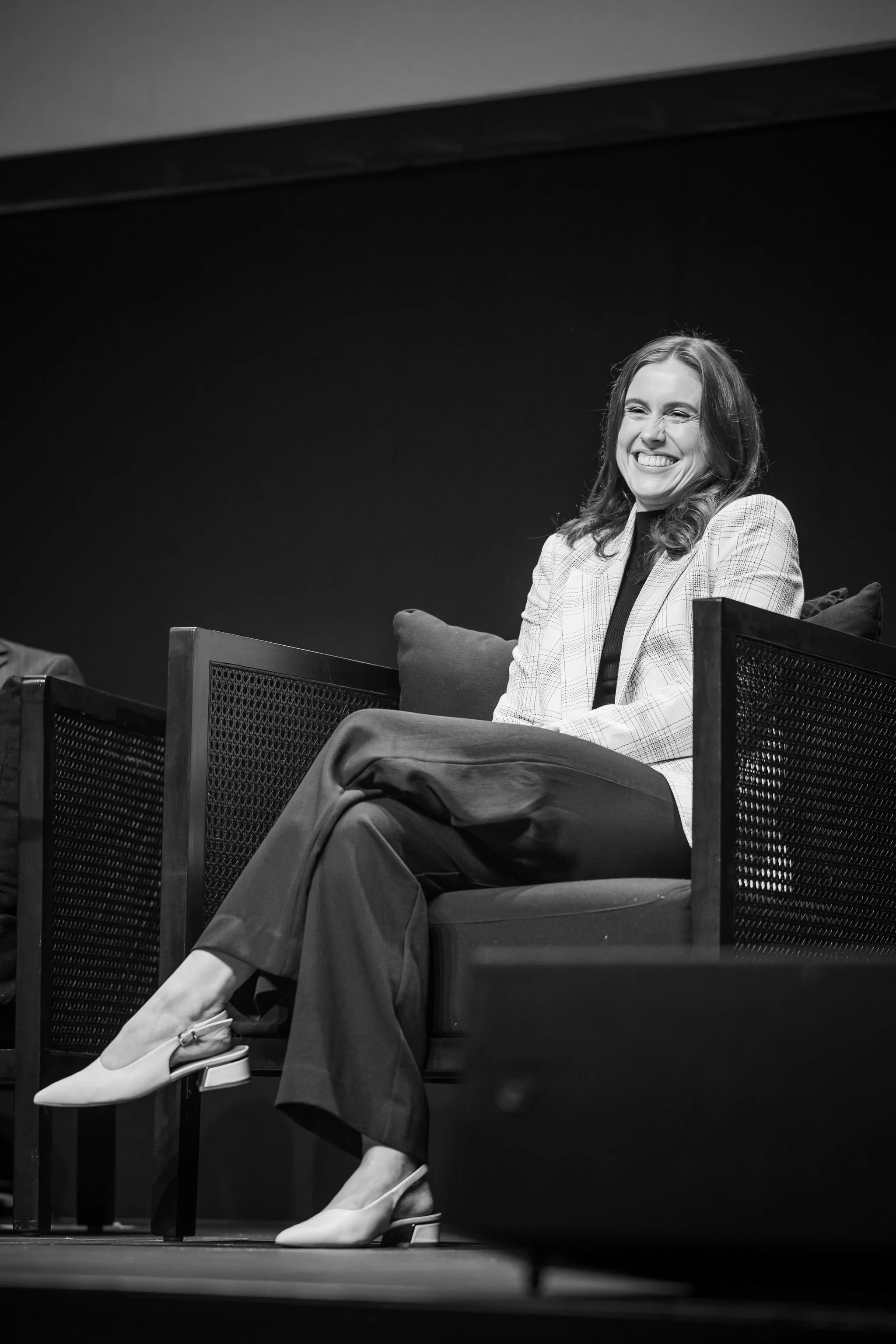 Black and white photo of a woman sitting on an armchair, smiling and laughing, wearing a plaid blazer, black turtleneck, and wide-leg pants, with her legs crossed, on a stage or panel discussion setting.