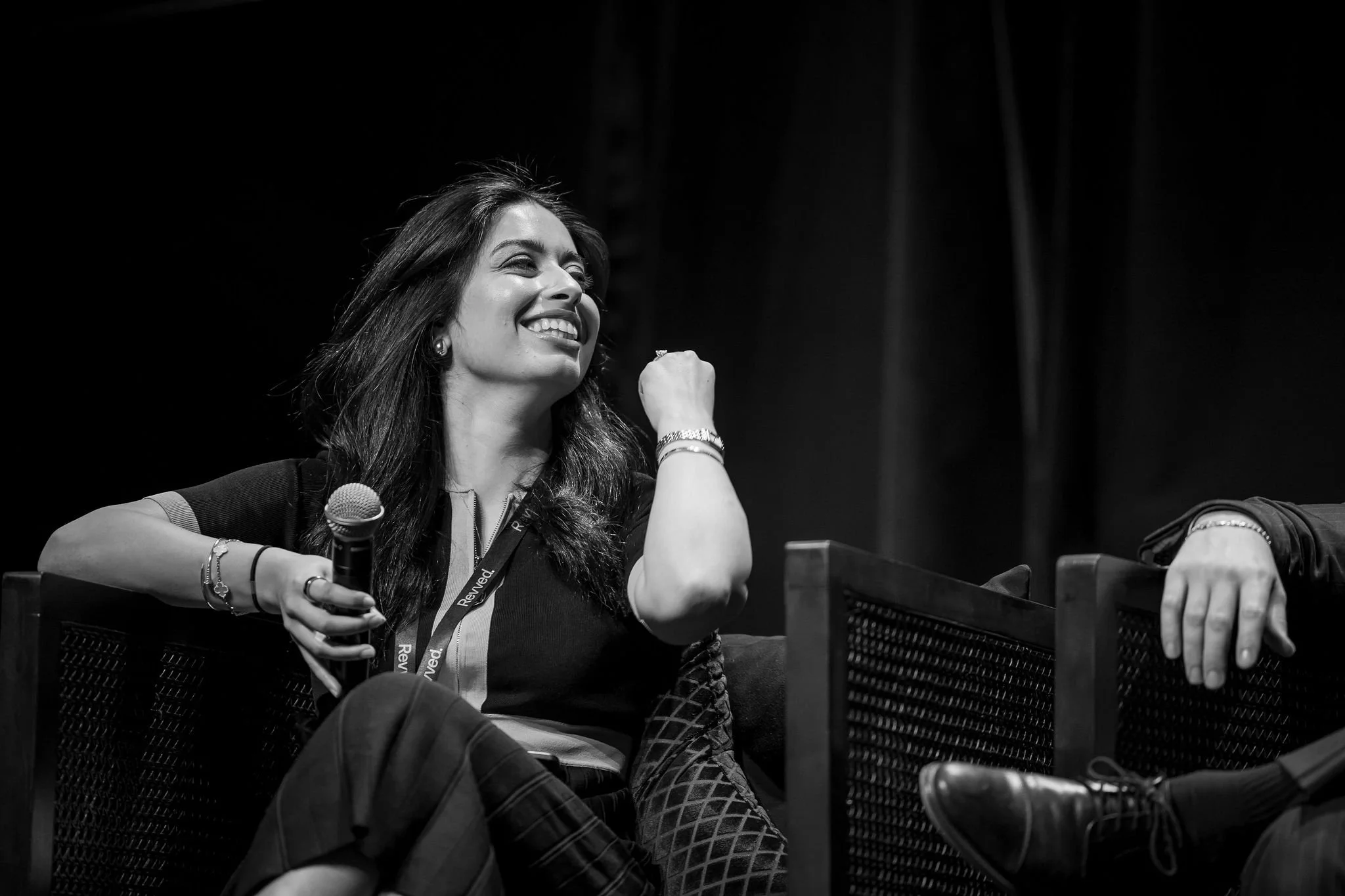 A woman sitting at a panel discussion, smiling, holding a microphone, with a logo on her shirt and a lanyard around her neck.