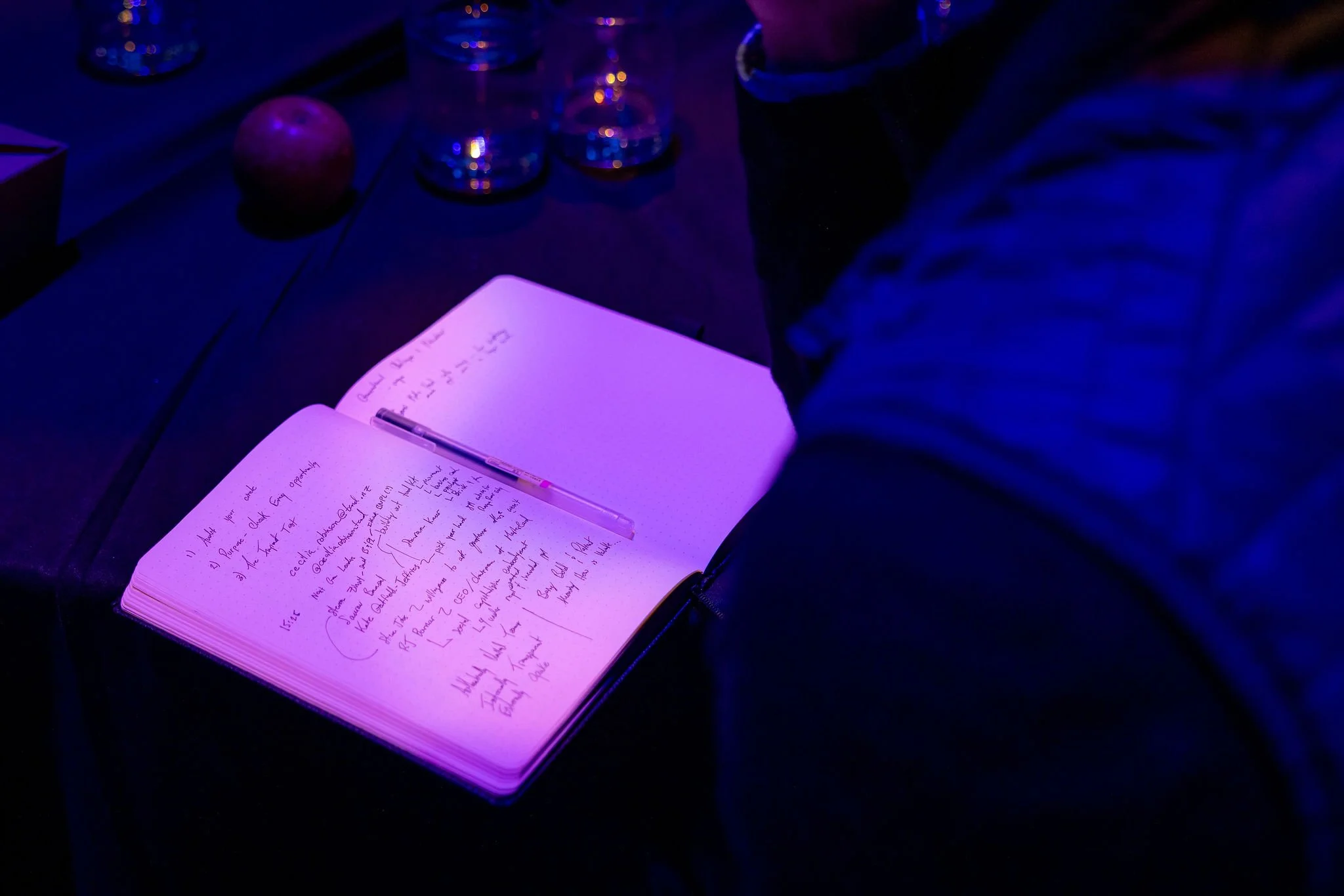 A notebook with handwritten notes is open on a dark surface illuminated by purple and blue lighting. Several glasses and an apple are visible in the background.
