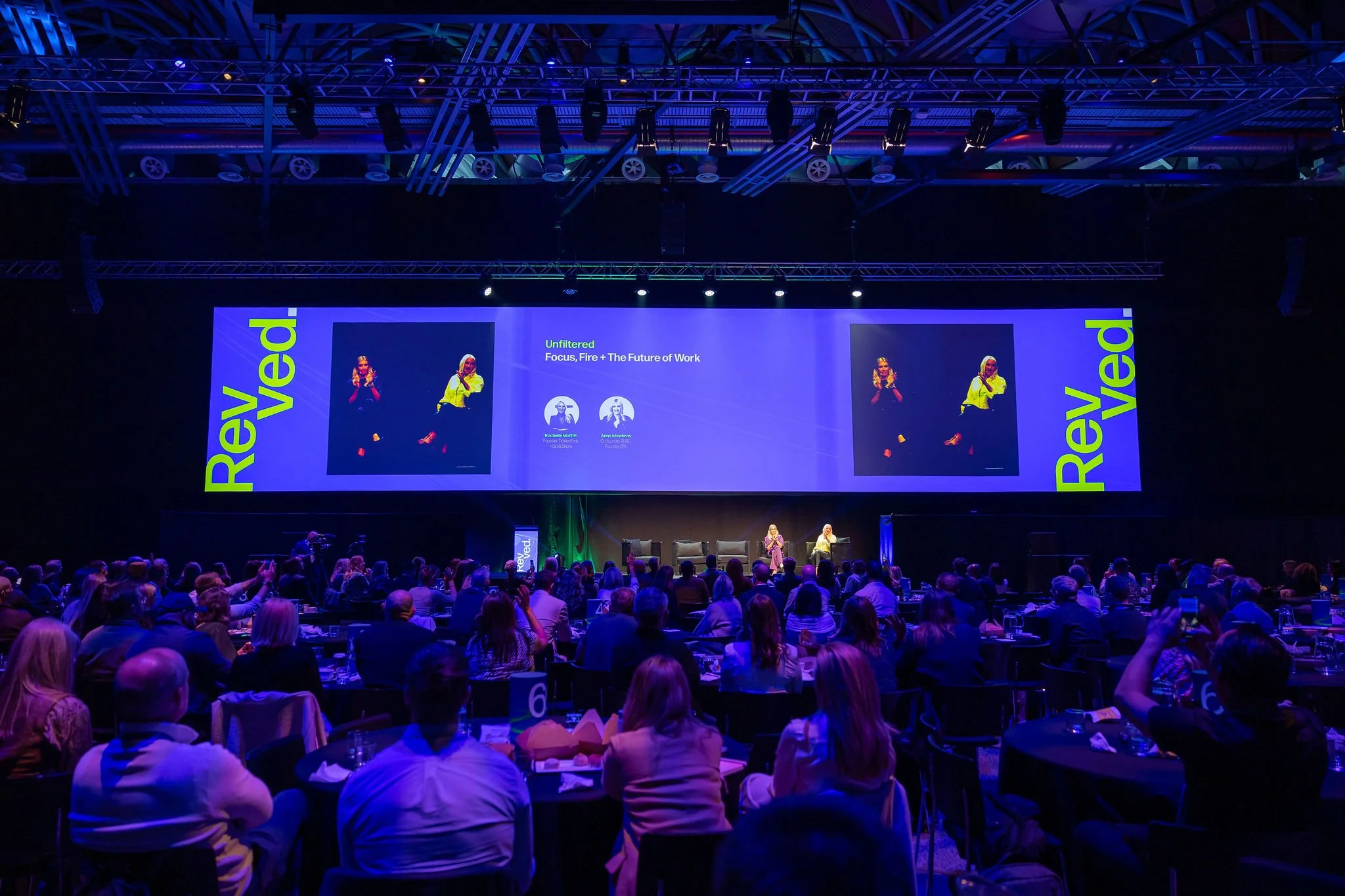 A large conference or event hall filled with people seated at round tables, watching a stage with a screen displaying the title 'Unfiltered: Focus, Fire + The Future of Work' and images of two women speakers.