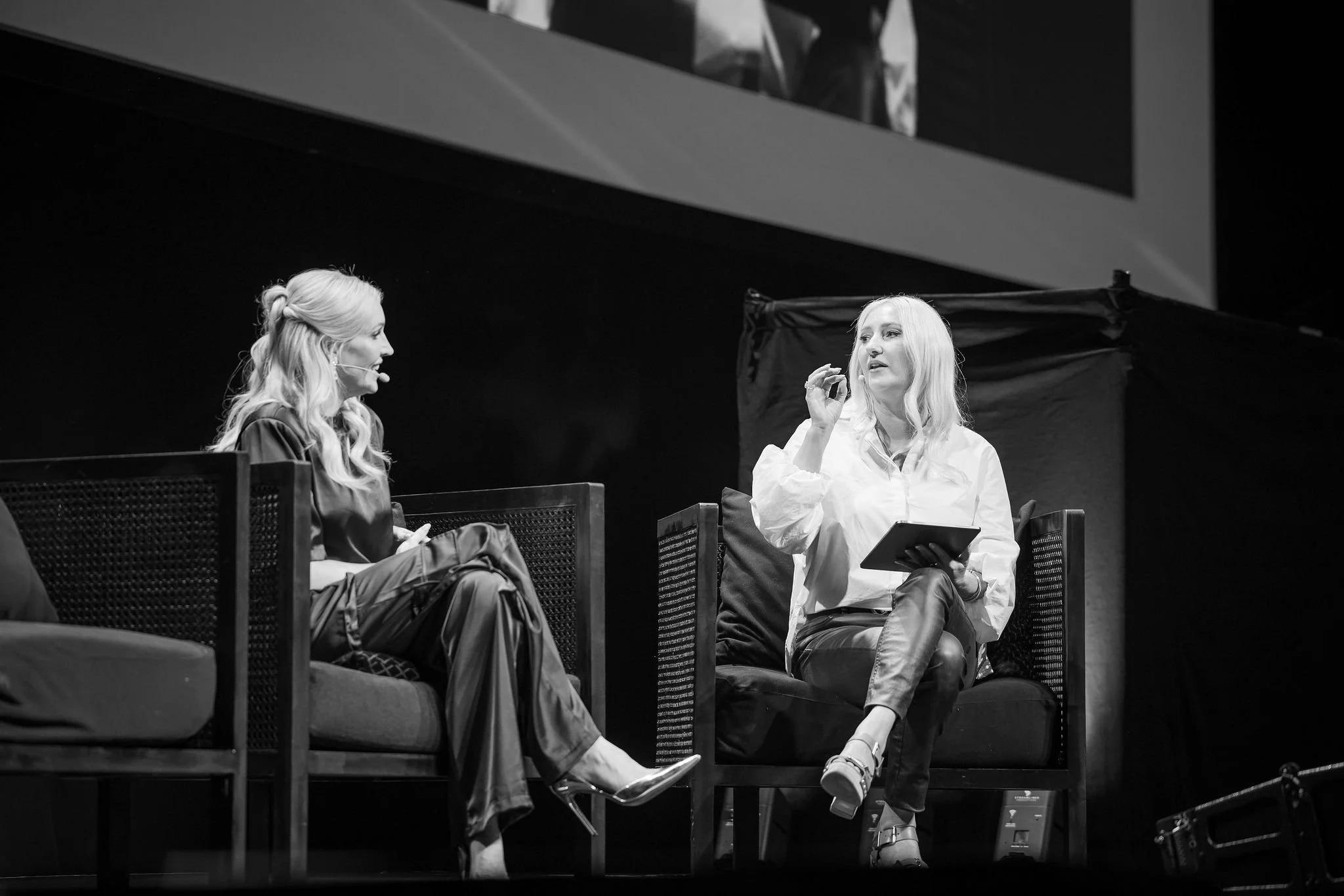 Two women with long, blonde hair sitting on a stage, engaging in conversation. One woman is wearing a dark jacket and pants, holding a phone, while the other woman is in a light-colored shirt and dark pants, holding a tablet. They are seated on bench