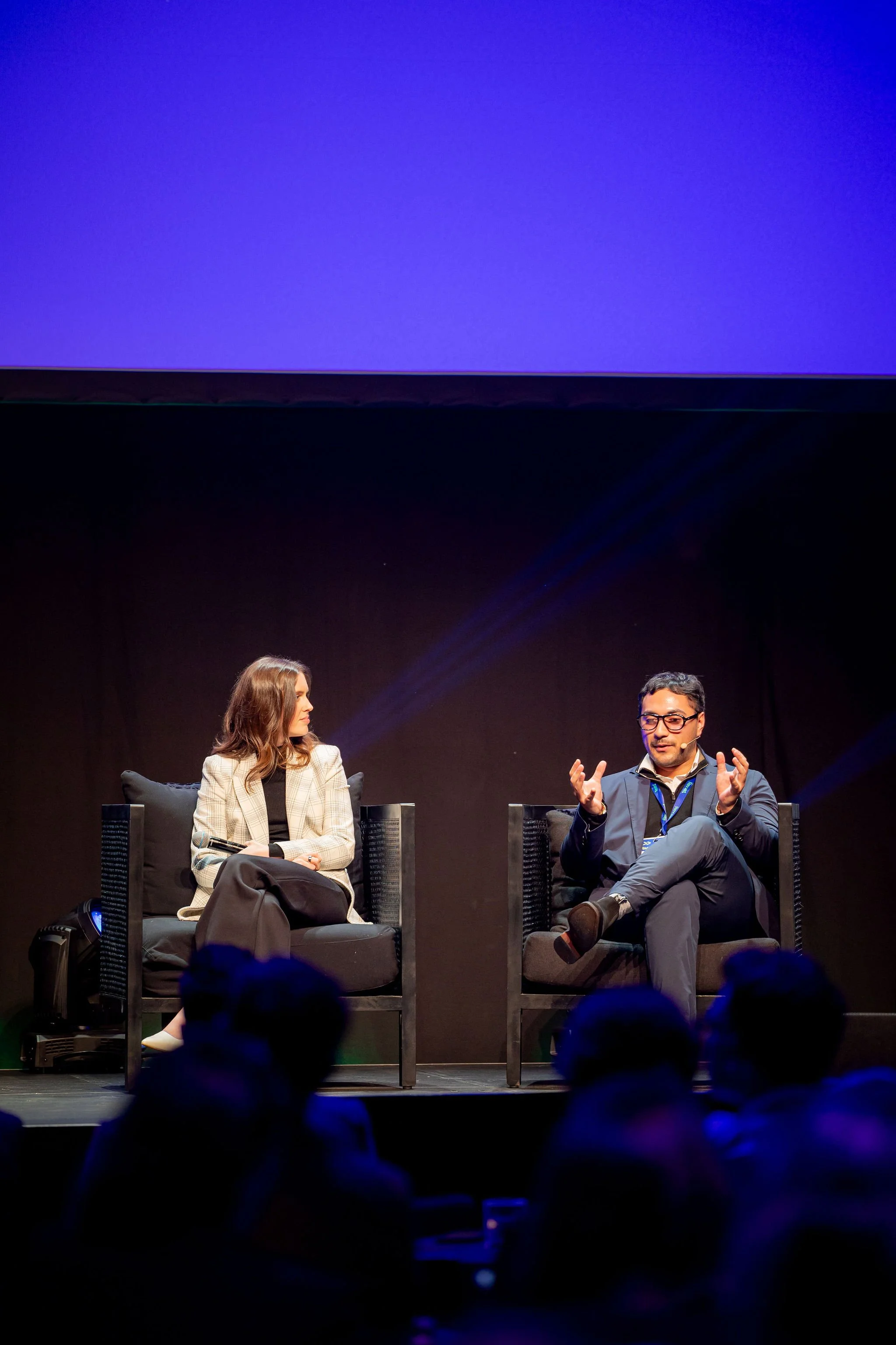 Two people are sitting on stage in black chairs during a panel discussion, with a large blue screen above them and audience members in the foreground.