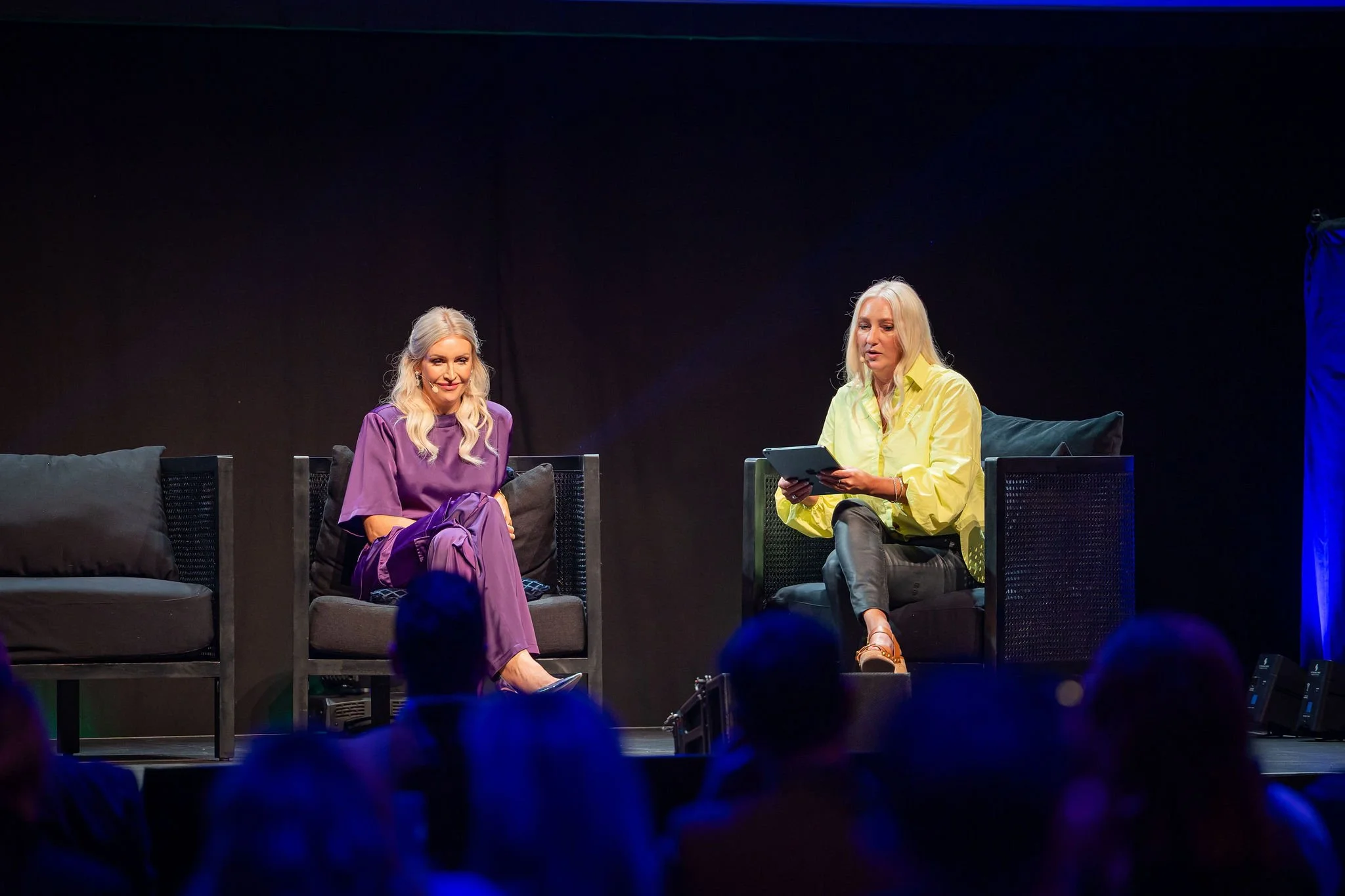 Two women sitting on a stage, engaged in a discussion. The woman on the left is wearing a purple outfit, and the woman on the right is wearing a yellow shirt, holding a tablet. There are black couches and a dark background.