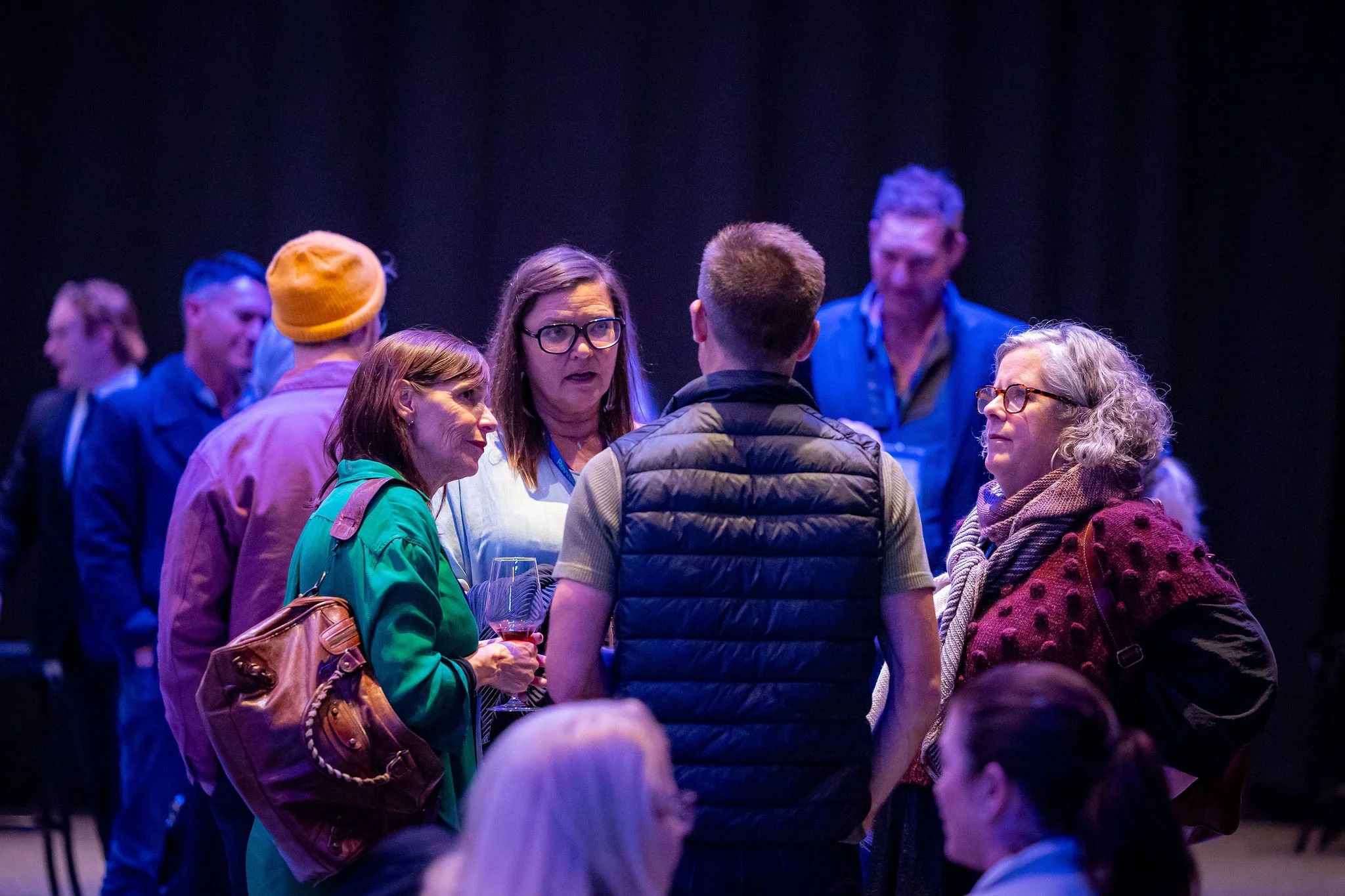 Group of people socializing at an indoor event with dark background, some holding drinks, engaged in conversation.