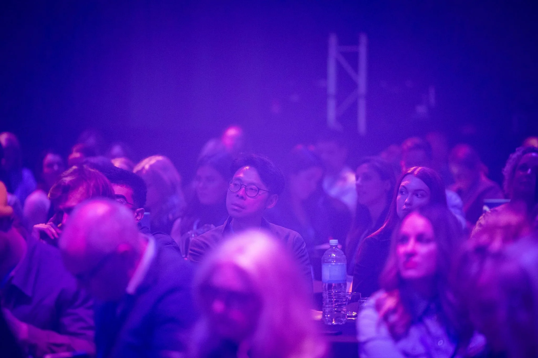 People attending the Revved conference under purple and blue lighting, seated and listening attentively, with one person in the foreground wearing glasses and a dark jacket.