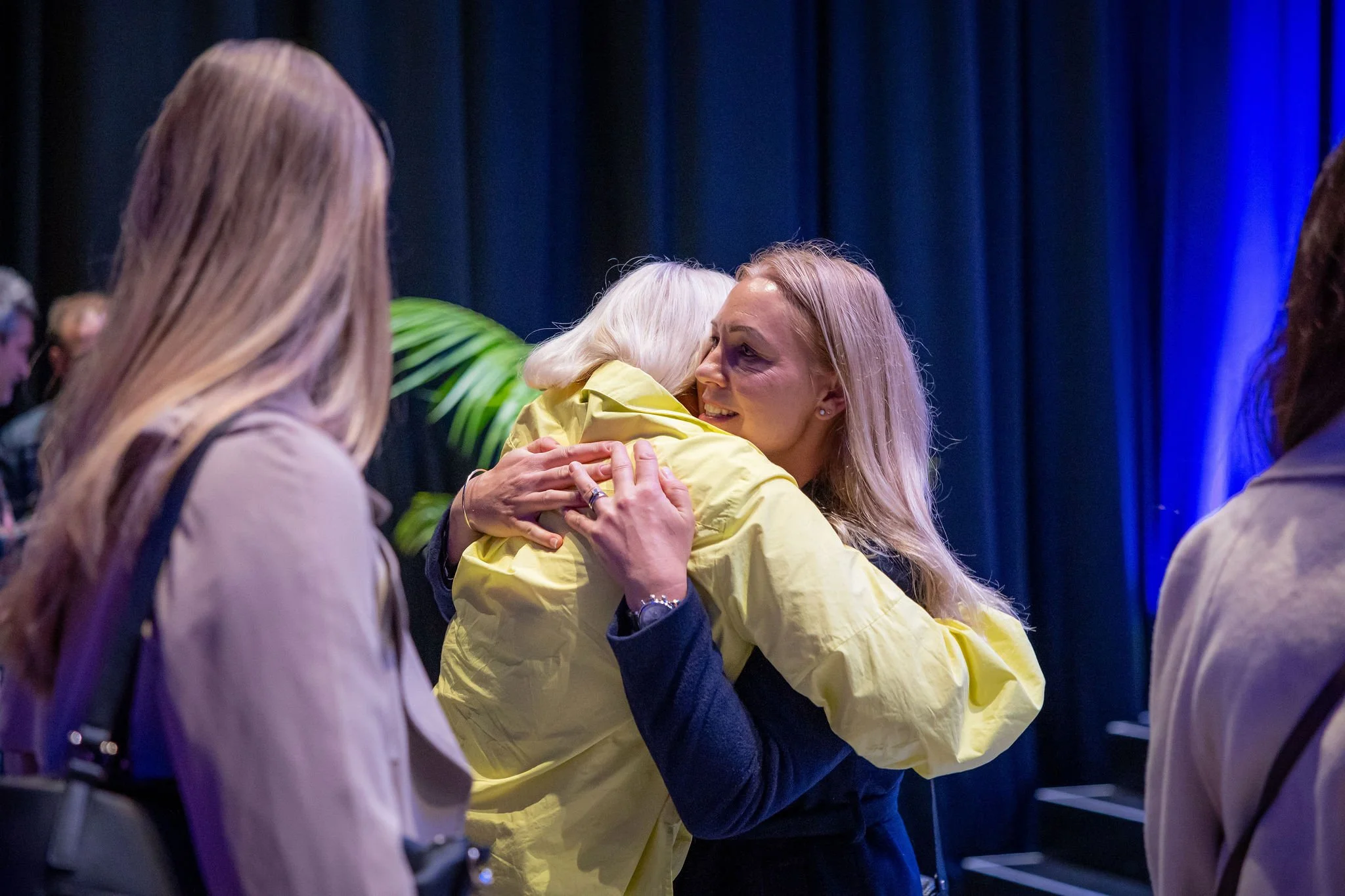 A group of women, with one woman hugging another woman in a yellow jacket, in an indoor setting with dark curtains and blue lighting.