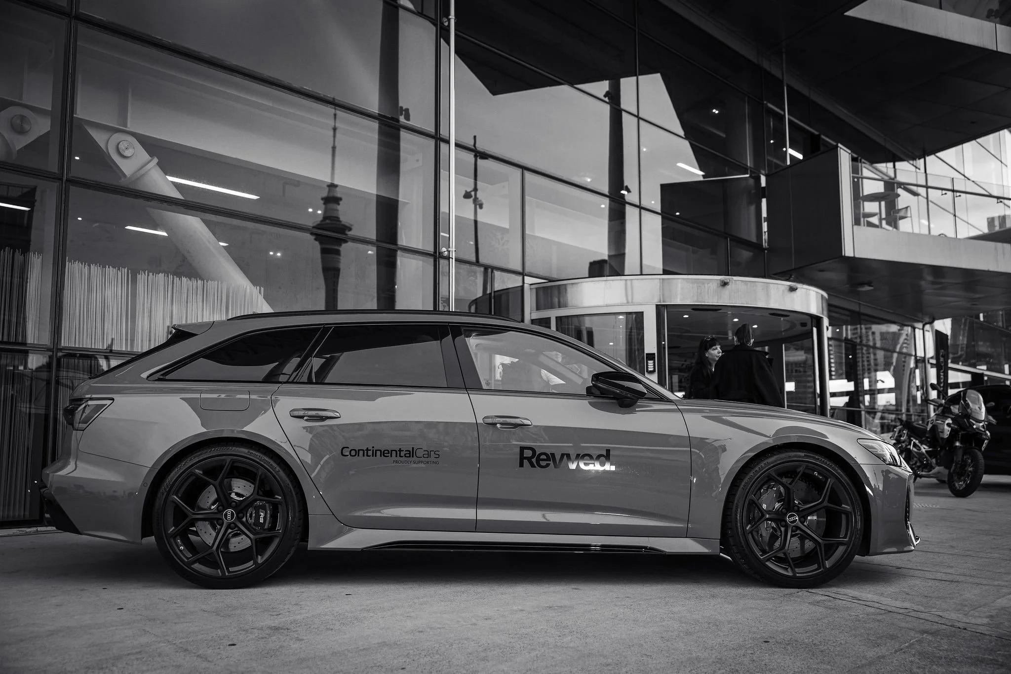 A silver sports car parked in front of a modern glass building with multiple stories and an overhang, with people visible in the background.