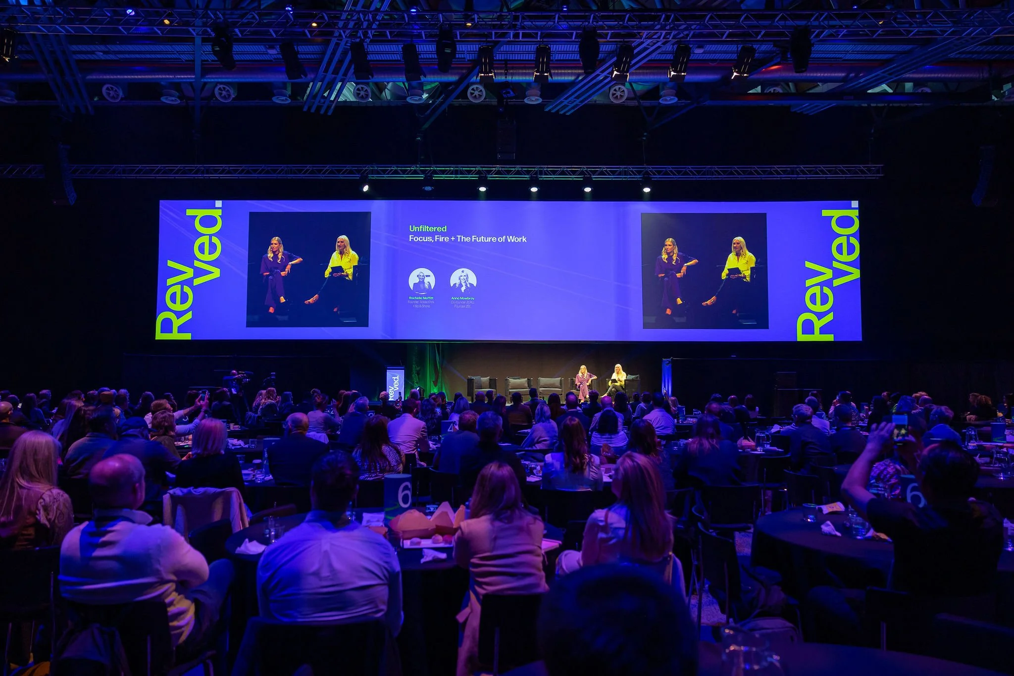 A large conference or event hall filled with people seated at round tables, facing a stage with two female speakers sitting and engaging in a discussion. The stage has a big screen displaying a presentation titled 'Unfiltered: Focus, Fire + The Futur