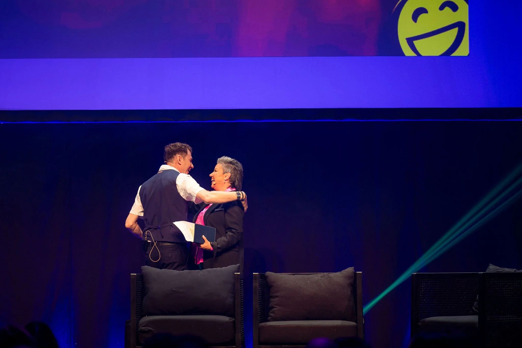 Two speakers smiling and embracing on stage during Revved, with purple and blue lighting and a large screen displaying a yellow smiling face in the background.