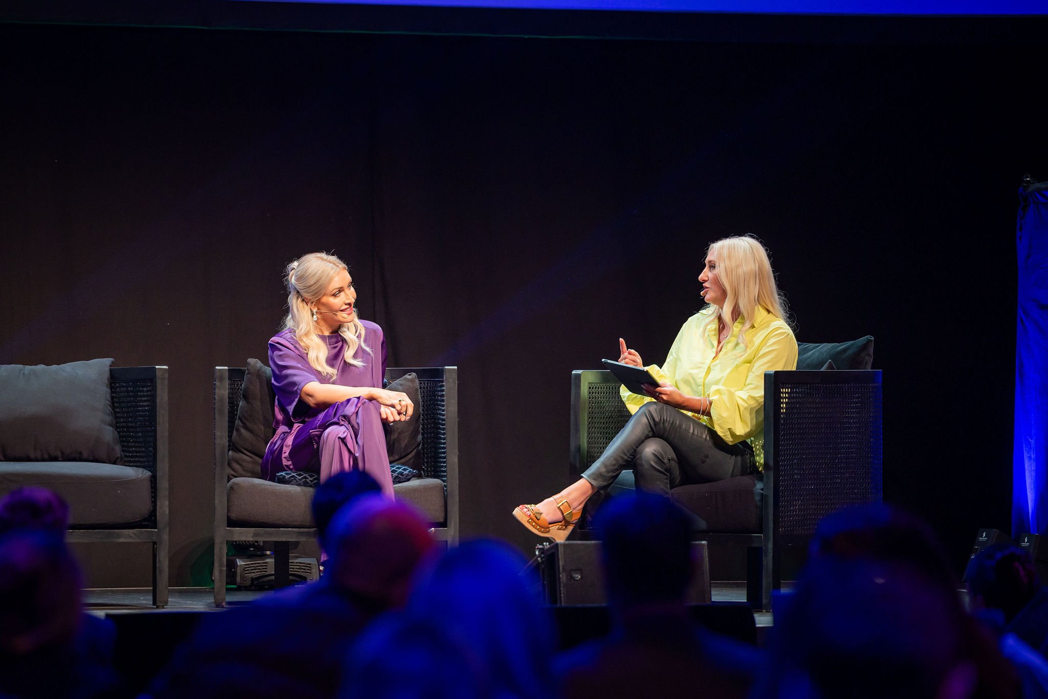 Two women sit on black wicker chairs with cushions on a stage, having a conversation. The woman on the left wears a purple dress, has blonde hair, and is smiling. The woman on the right wears a yellow blouse, black pants, and golden shoes, holding a 