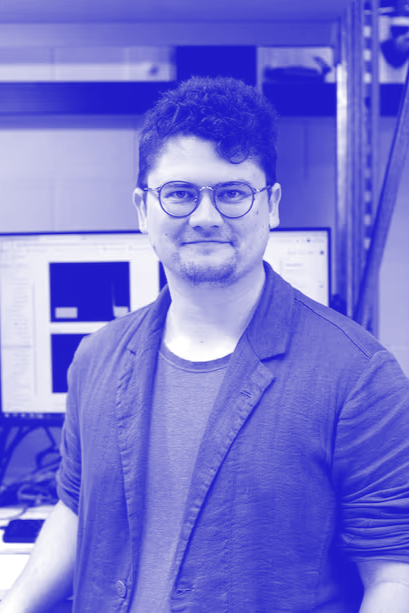 A man with curly hair and glasses standing in front of computer monitors in a server room or tech workspace.