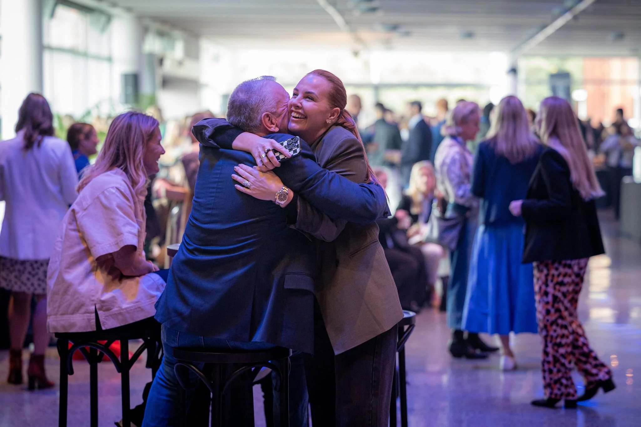 A woman and an older man hugging and smiling at an indoor social event with other people in the background.