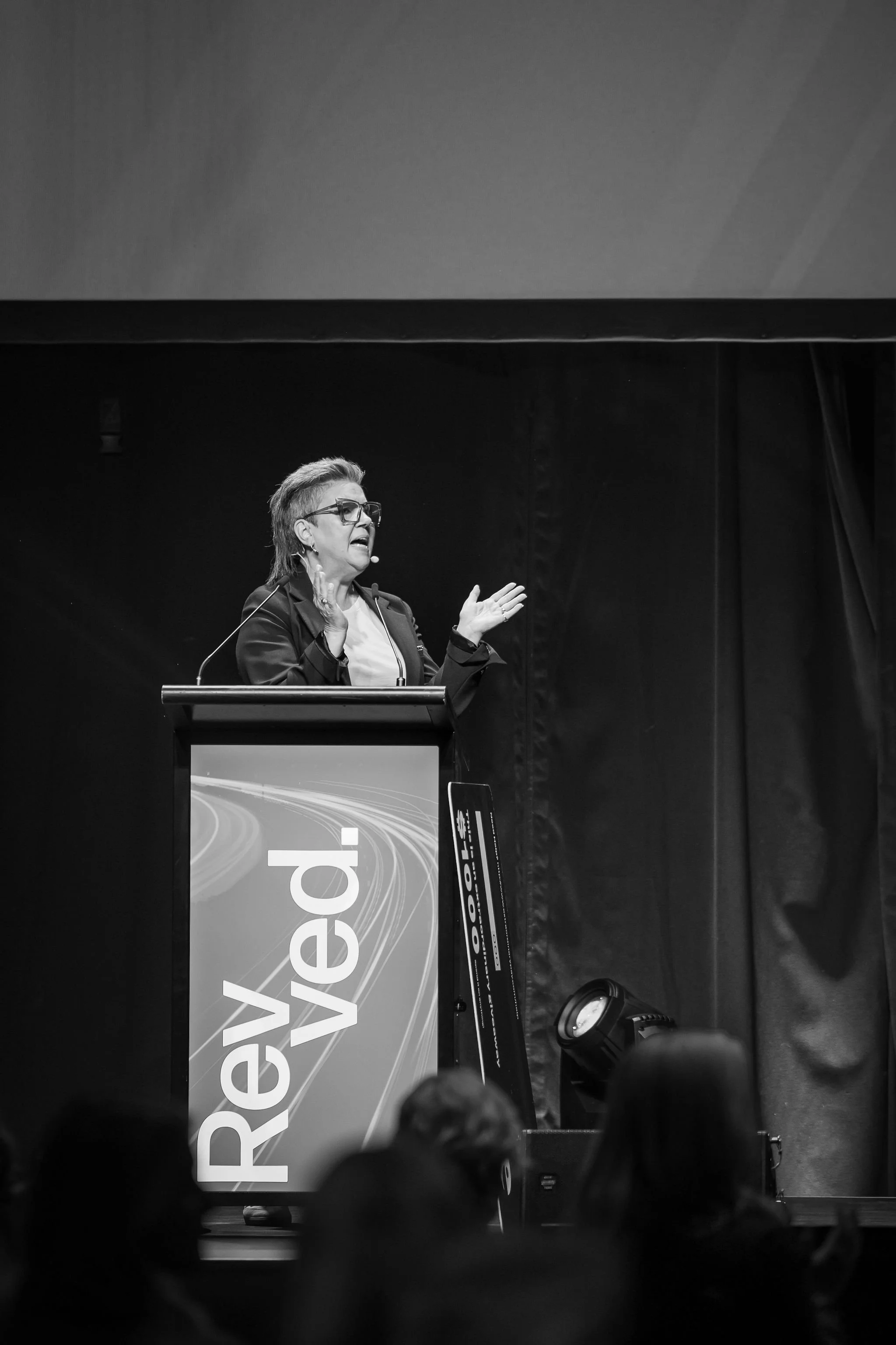 Black and white photo of a woman speaking at a podium with a digital display, with an audience in the foreground.