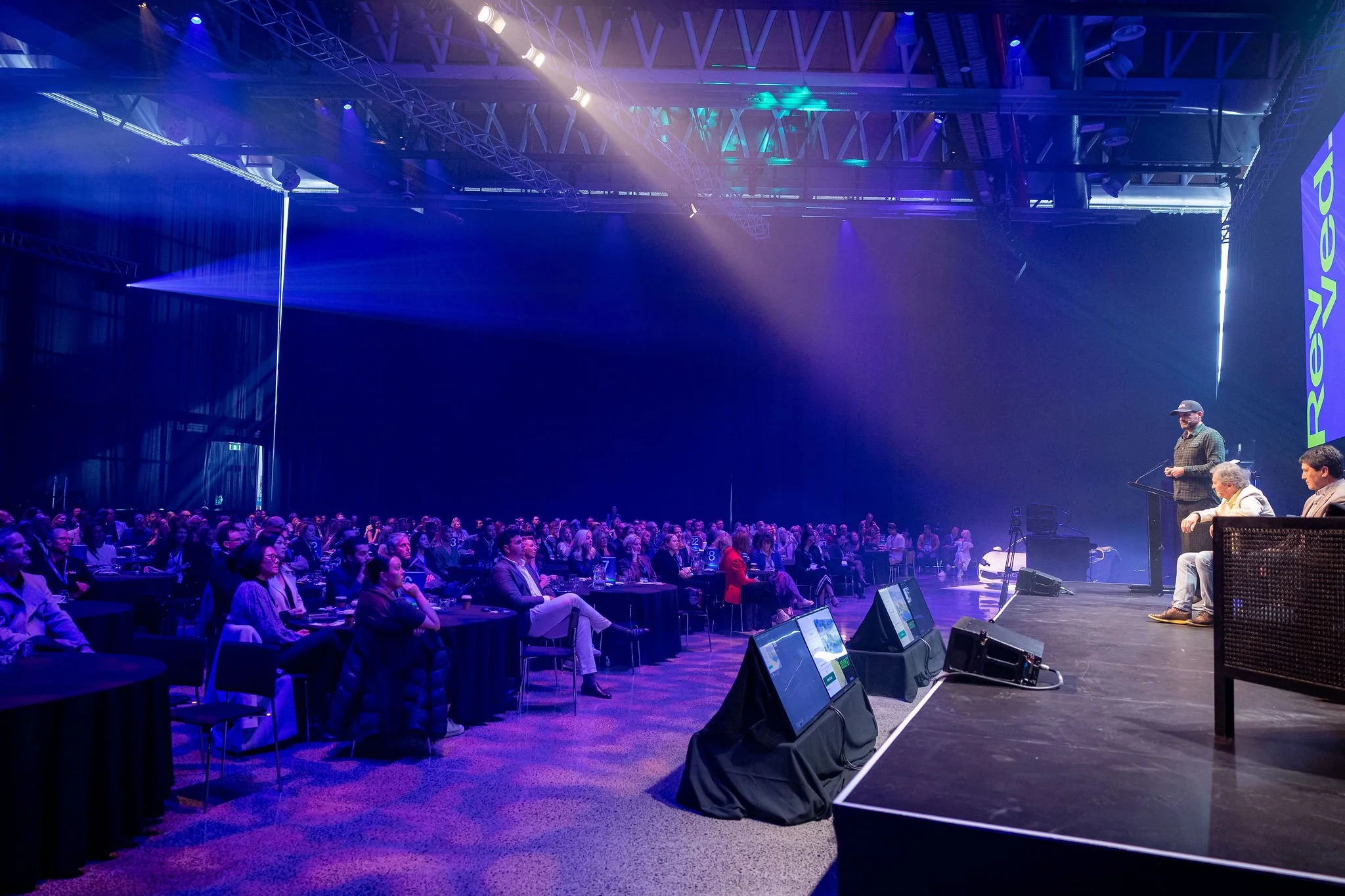 Revved conference with many attendees sitting at tables, facing the stage with panel of speakers, illuminated with colorful stage lighting and a screen displaying the word 'powered'.