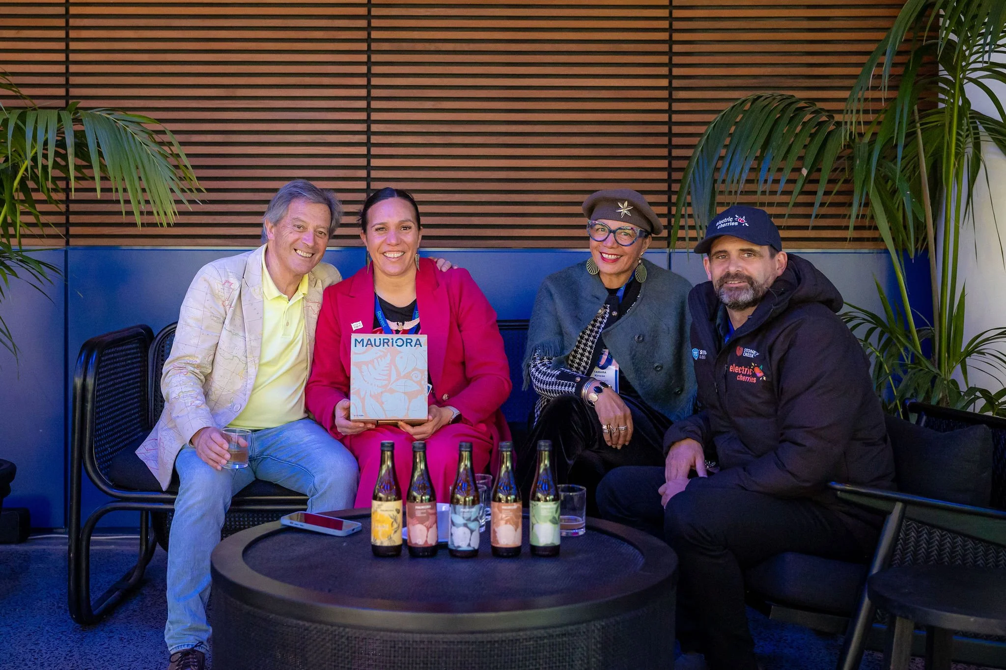 Four people sitting together at a table, smiling. The woman in the center is holding a booklet titled "MAURORA." There are six bottles on the table in front of them, and a man on the far right is wearing a shirt with a logo that says "electric cherri