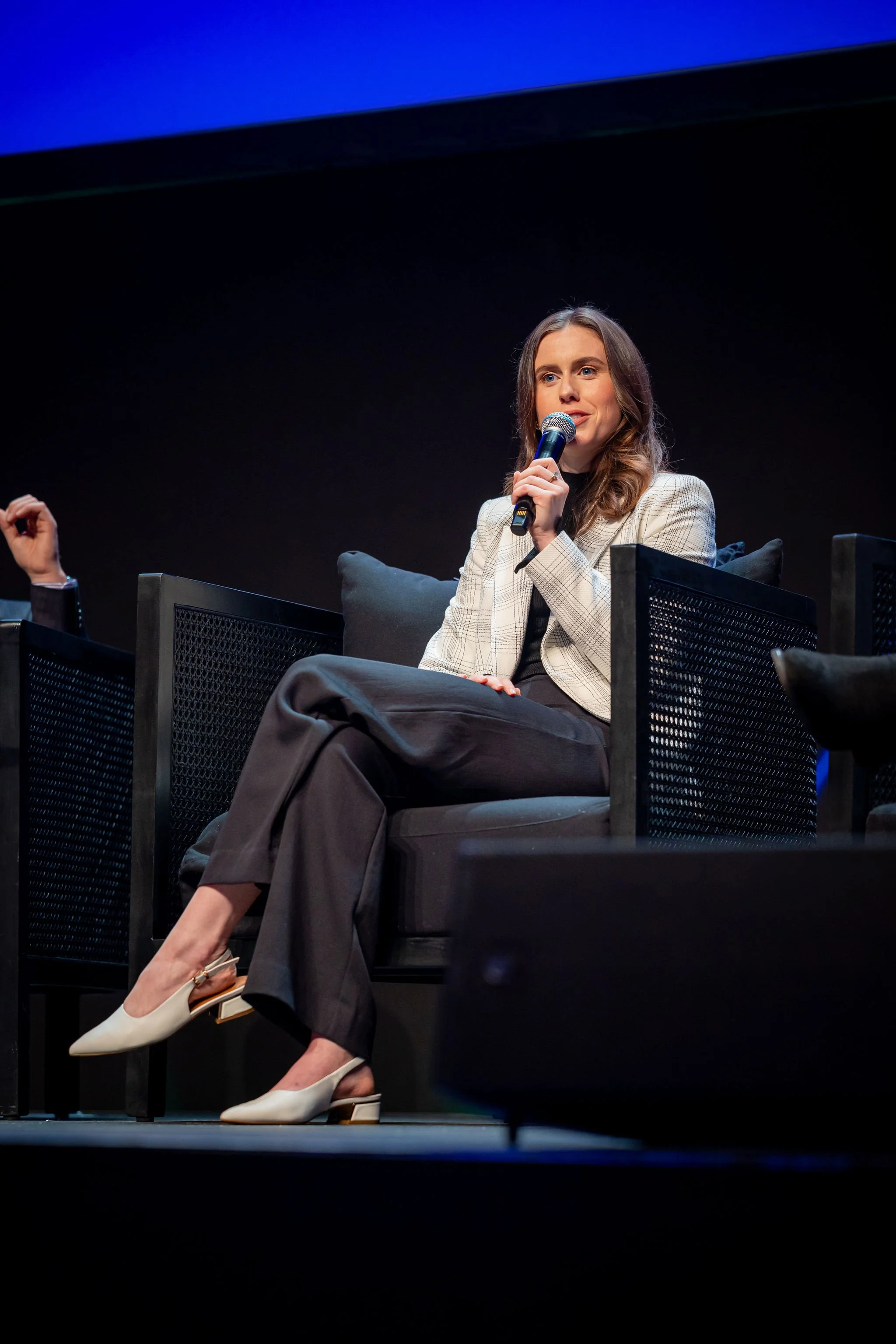 A woman with long brown hair wearing a beige blazer and black pants sitting on stage with a microphone in her hand during a panel discussion.