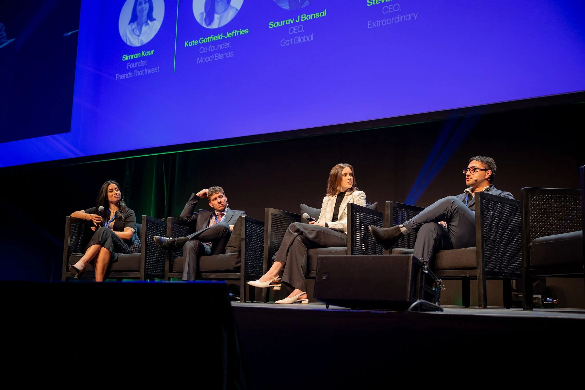 A panel discussion at a conference with four people seated on stage. A large screen behind them displays pictures and names of the speakers, including Simran Kaur, Kate Gaffield Jeffries, Saurav J Bansal, and Steve. The stage is dark, and the panelis