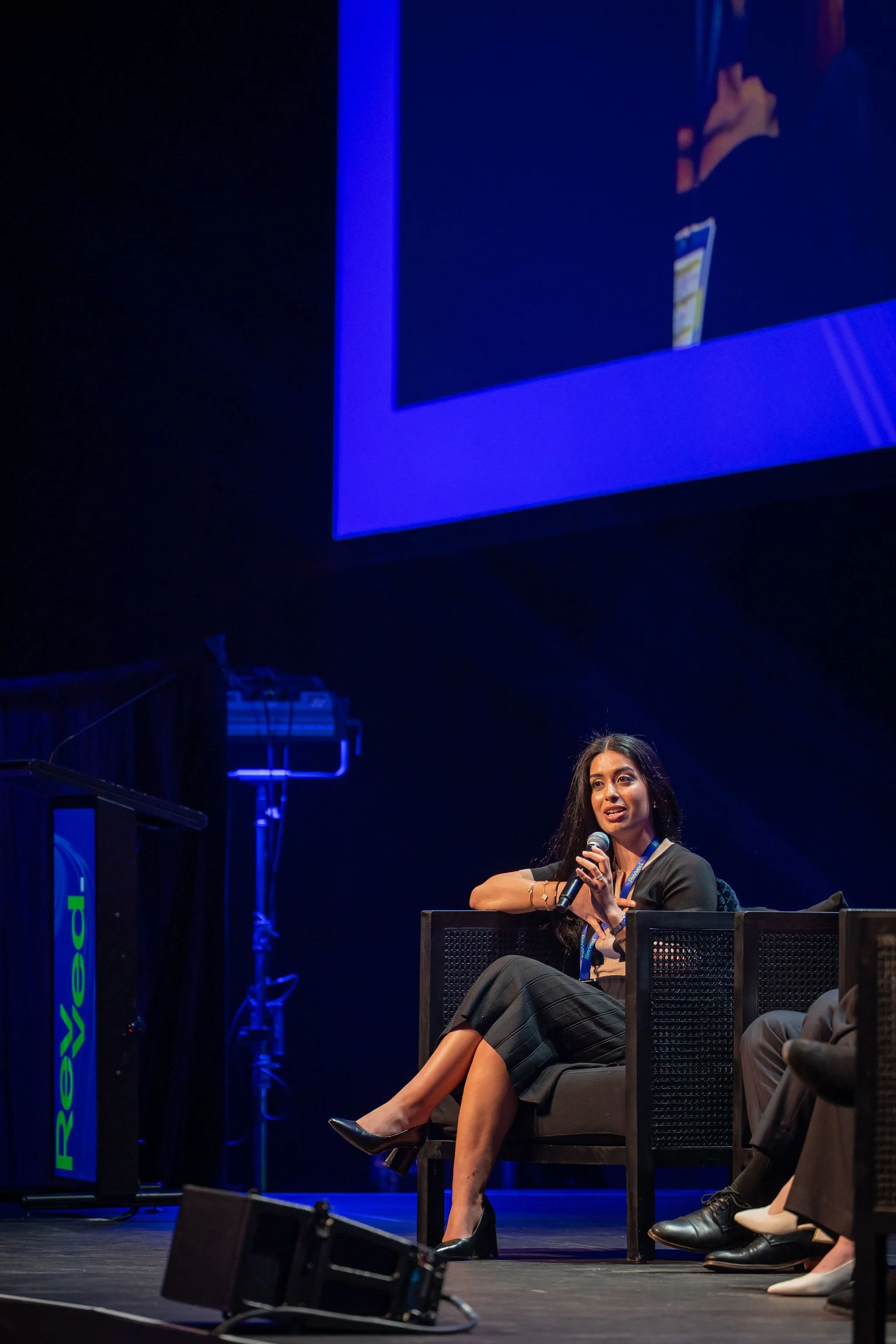 A woman sitting on a chair on stage, holding a microphone, participating in a panel discussion or conference, with other seated individuals nearby, and a large screen behind her displaying part of her image.