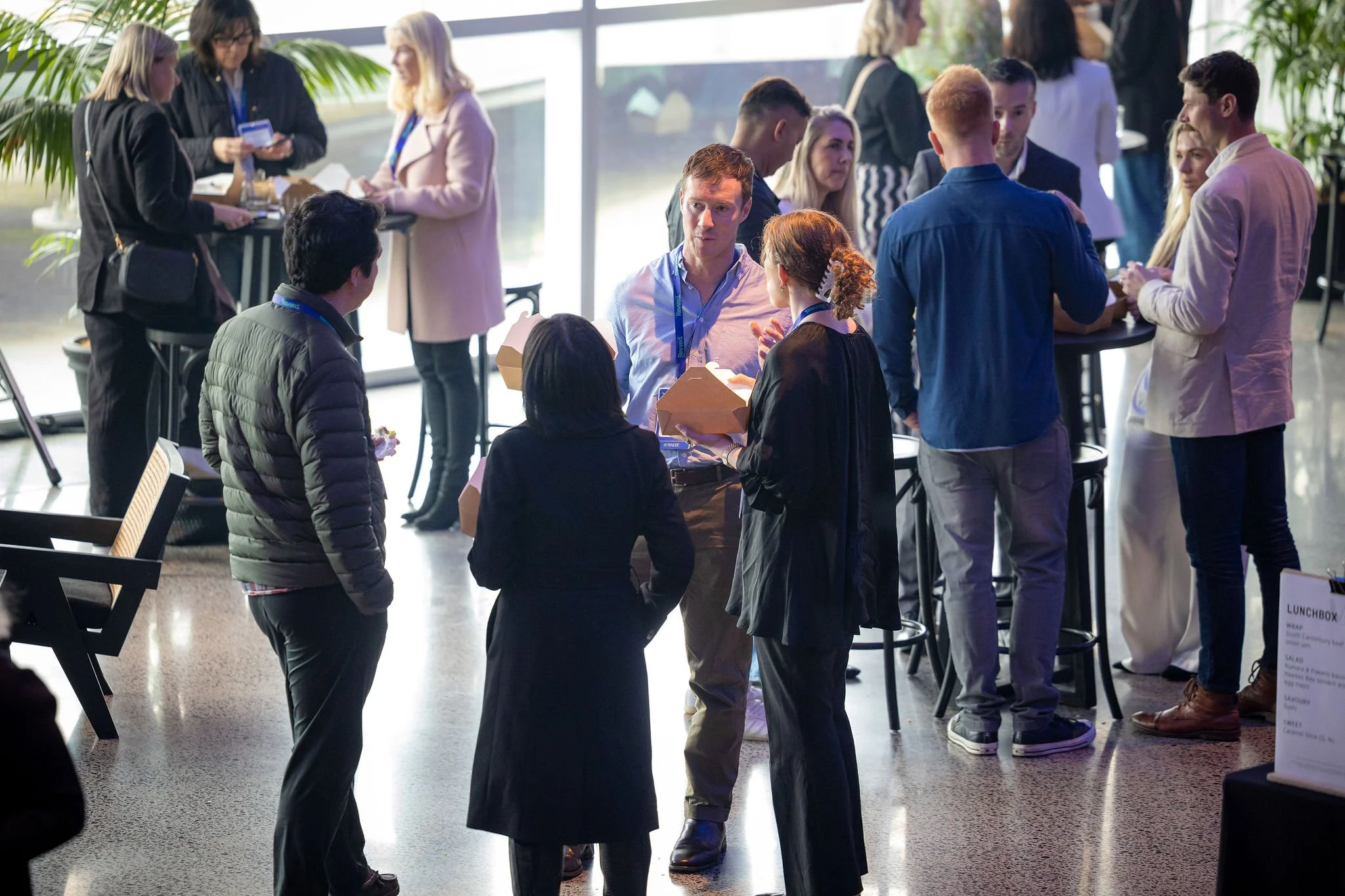 Group of people at a social event or conference, standing and conversing in a well-lit space with large windows and plants.