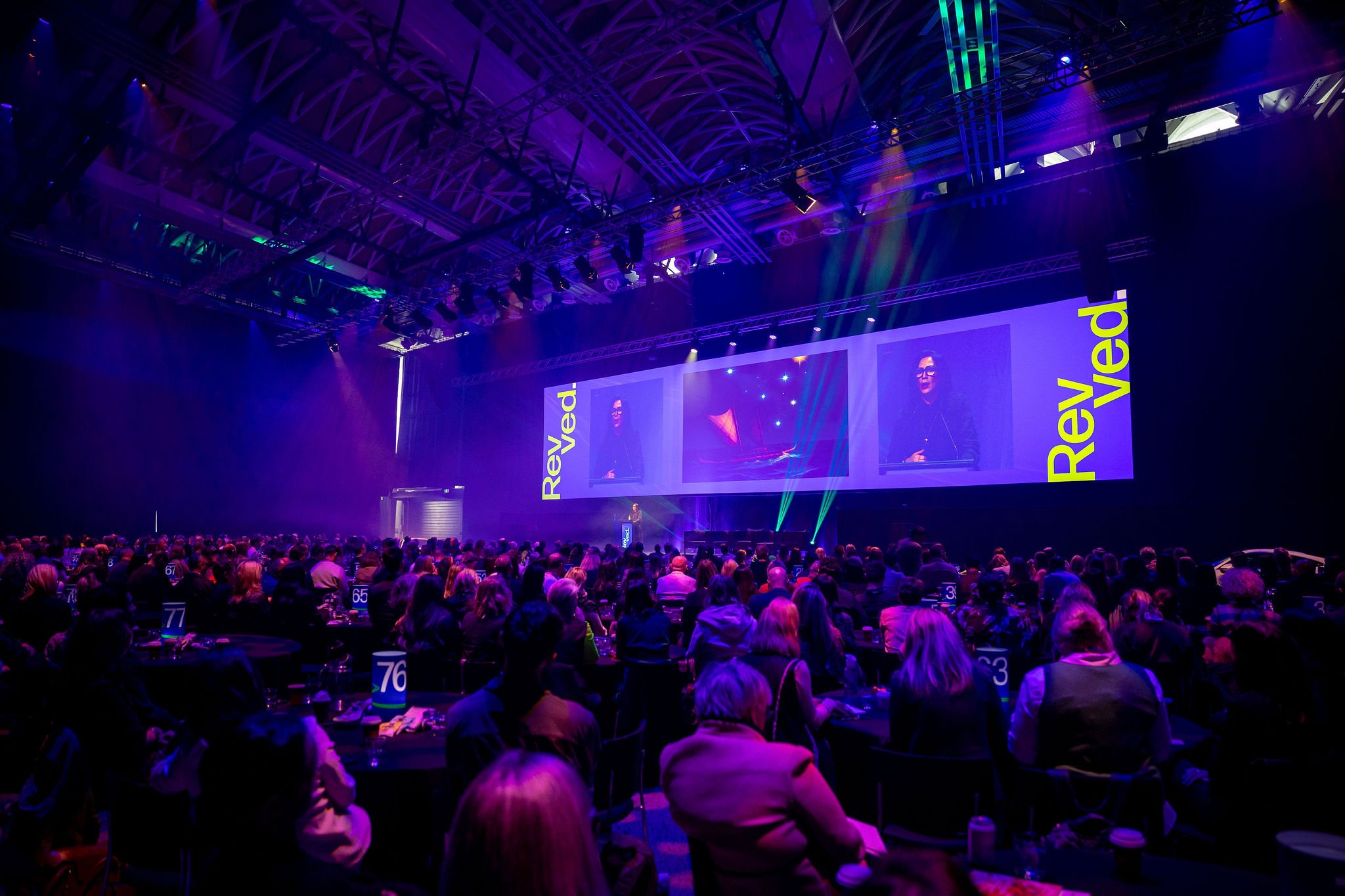 Revved conference auditorium filled with people, with a stage at the front displaying multiple screens and the words 'Rev 3.0' in yellow on a purple background. 