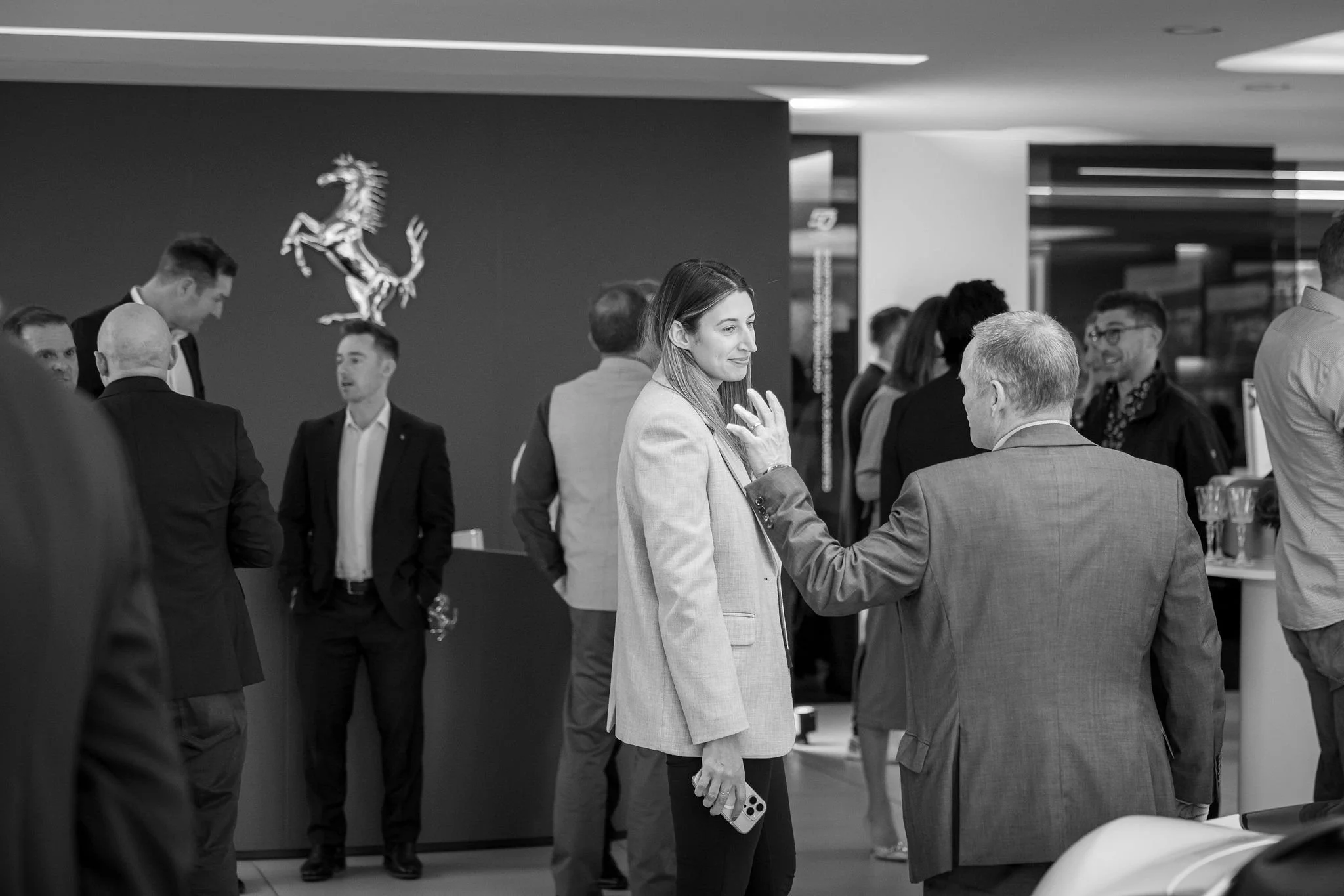 A group of people in a professional setting, engaging in conversations, with a woman in a blazer holding a mobile phone in her right hand and wearing glasses on the front of her shirt, smiling and appearing to be in a discussion.