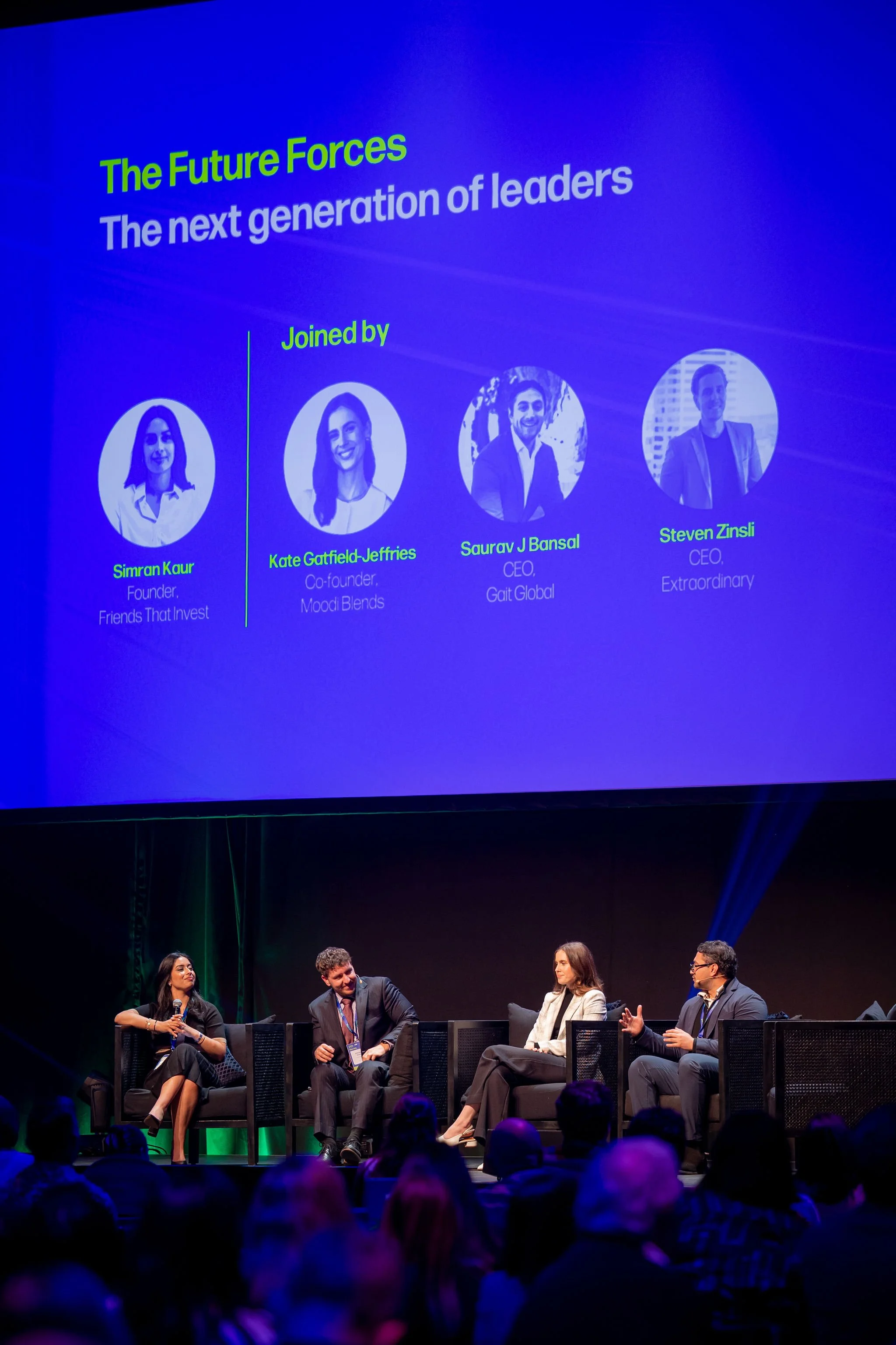 Panel discussion at a conference with five panelists seated on a stage, four men and one woman, with a large screen displaying names and titles behind them. Audience silhouettes are visible in the foreground.