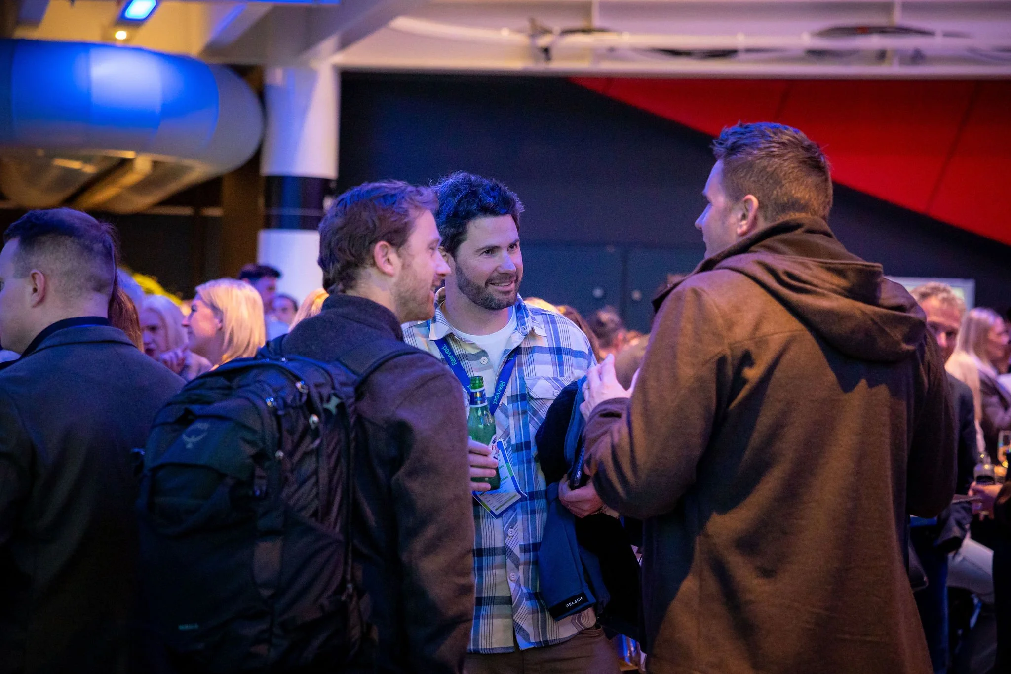 Three men are engaged in conversation at a crowded indoor event. The man in the middle is wearing a plaid shirt and holding a bottled beverage, while the other two men are facing him and listening. Background shows other attendees and vibrant lightin