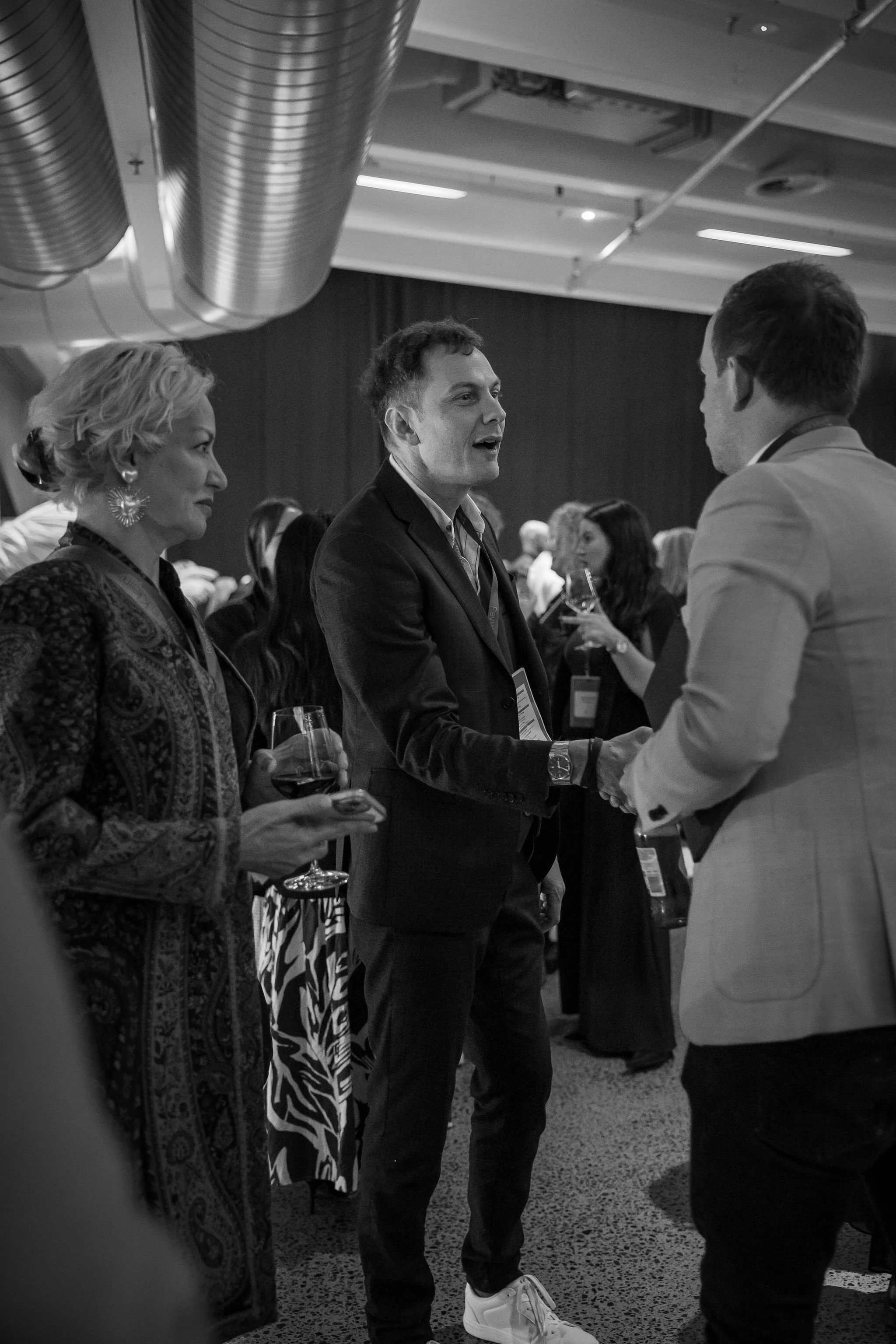 Three people are engaged in conversation at an indoor event. The man in the center is shaking hands with the man on the right. The woman on the left holds a glass of wine and a phone. The setting appears to be professional or networking event with ot