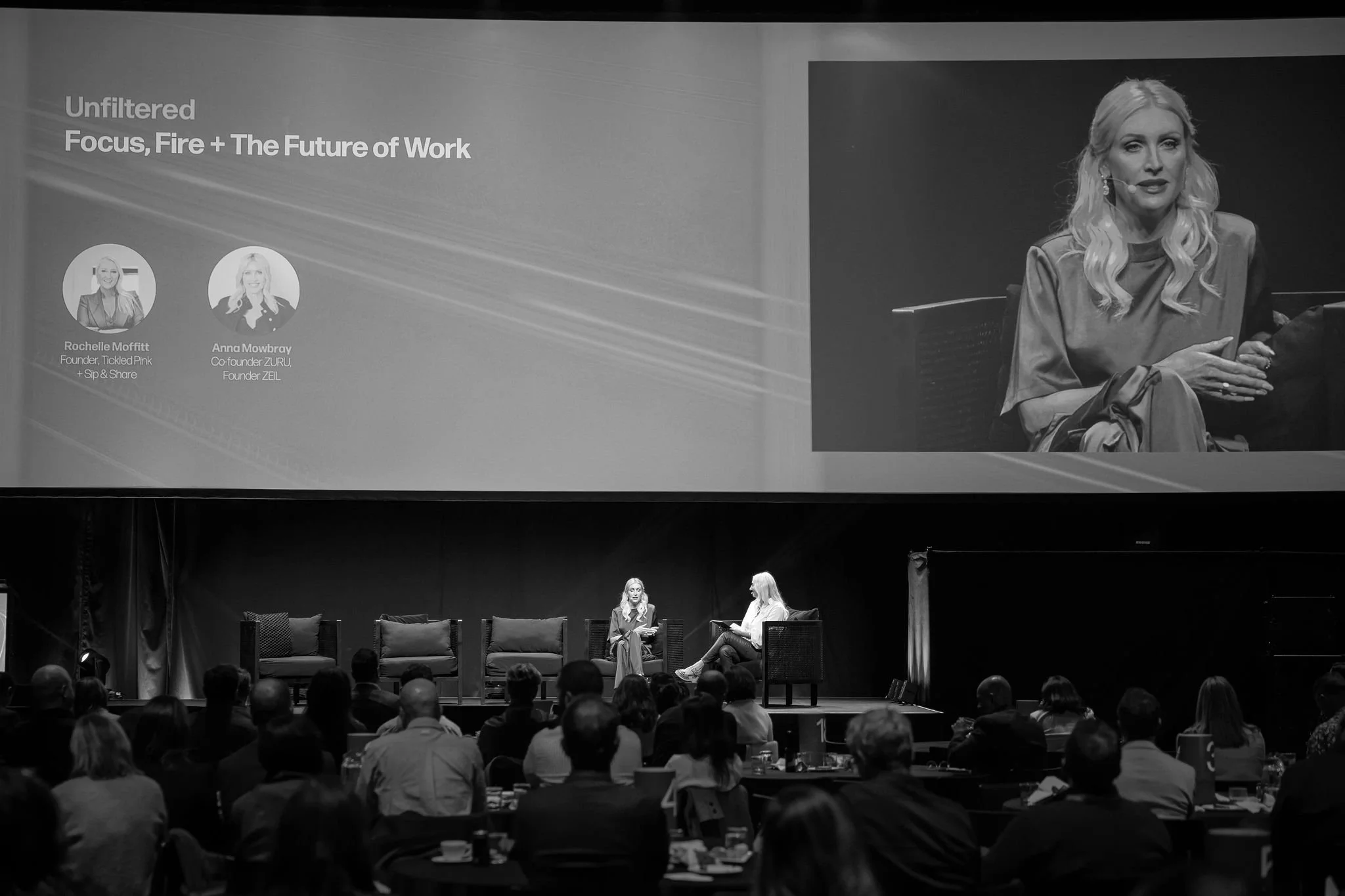 A panel discussion at a conference with two women seated on stage and a large screen displaying their names and titles. Audience members are visible in the foreground.